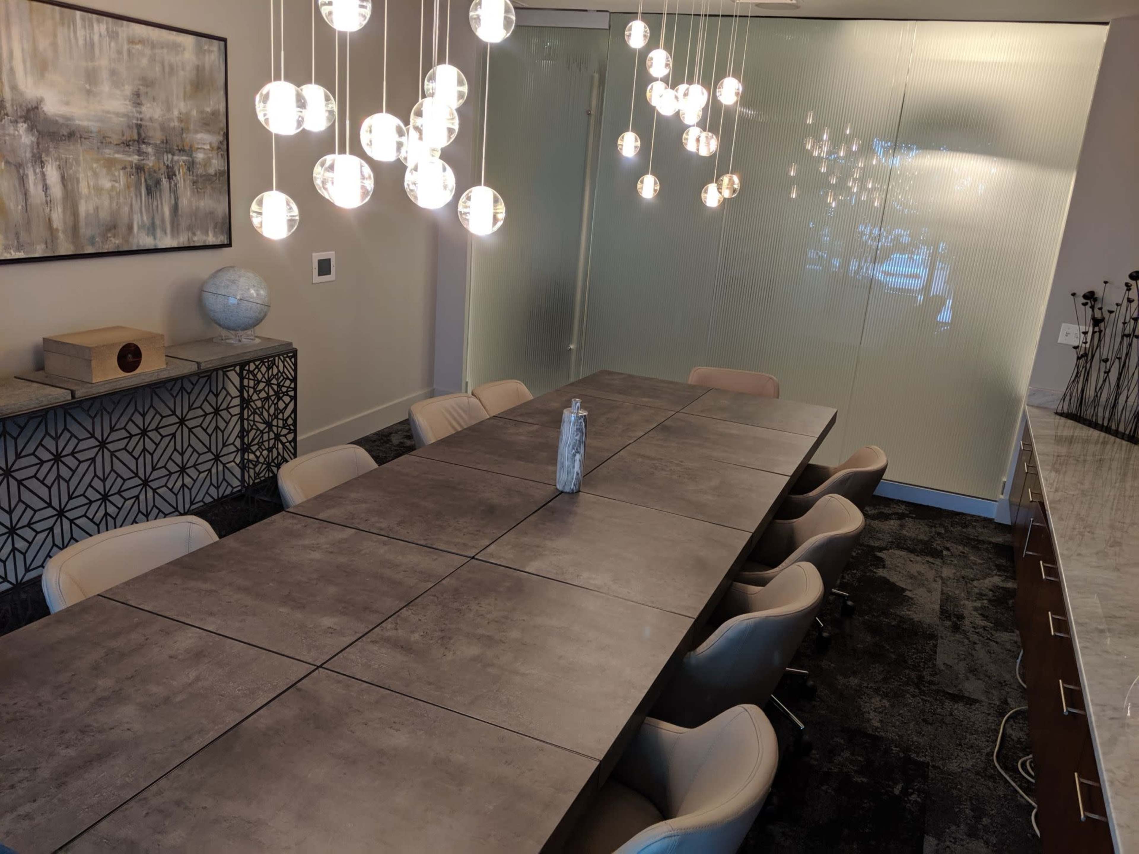 A modern conference room with a long concrete table, ten beige chairs, pendant lights above, and a frosted glass wall in the background.