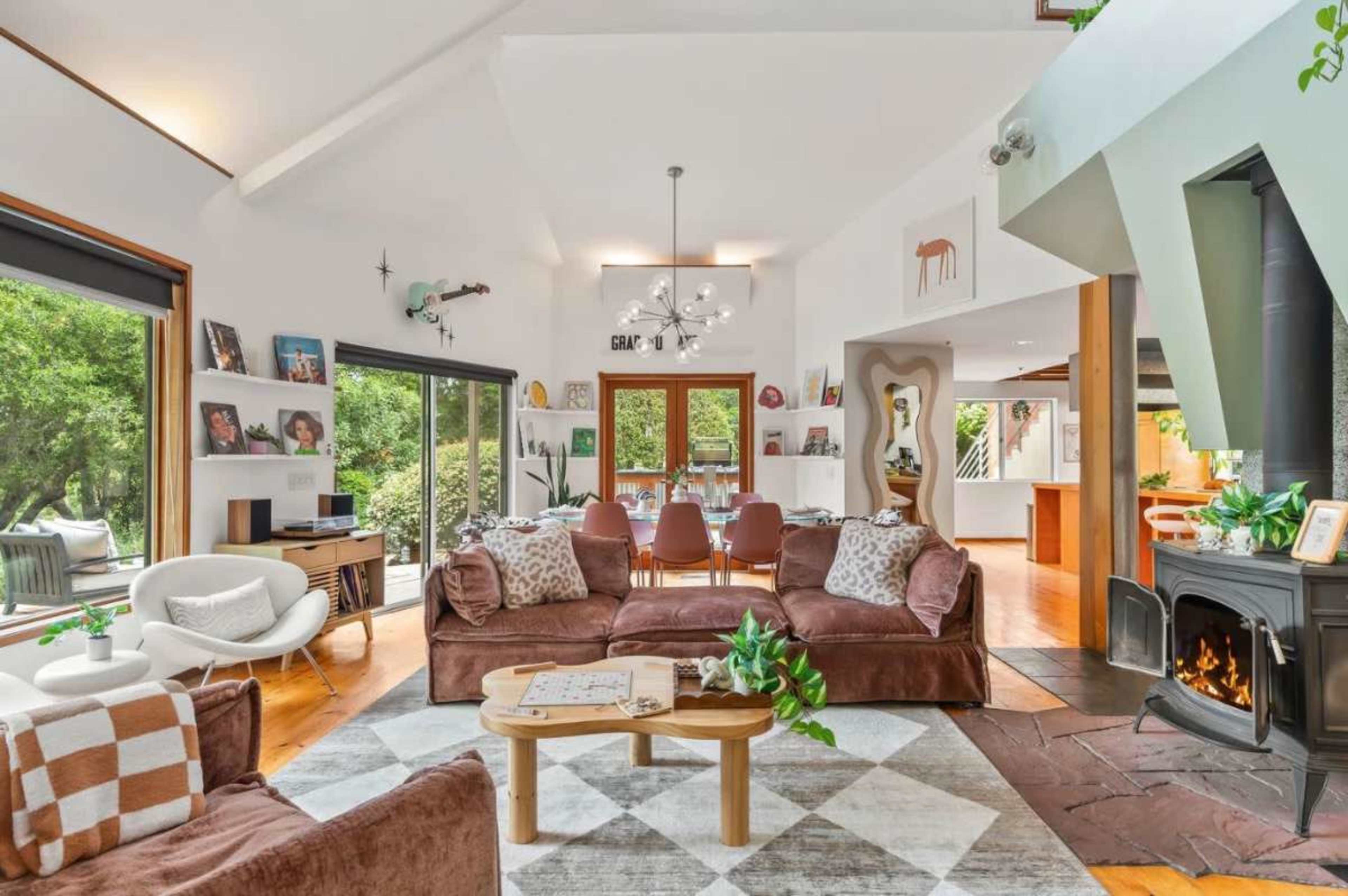 The image shows a cozy living room with brown sofas, a coffee table on a patterned rug, and large windows that overlook a green outdoor area.
