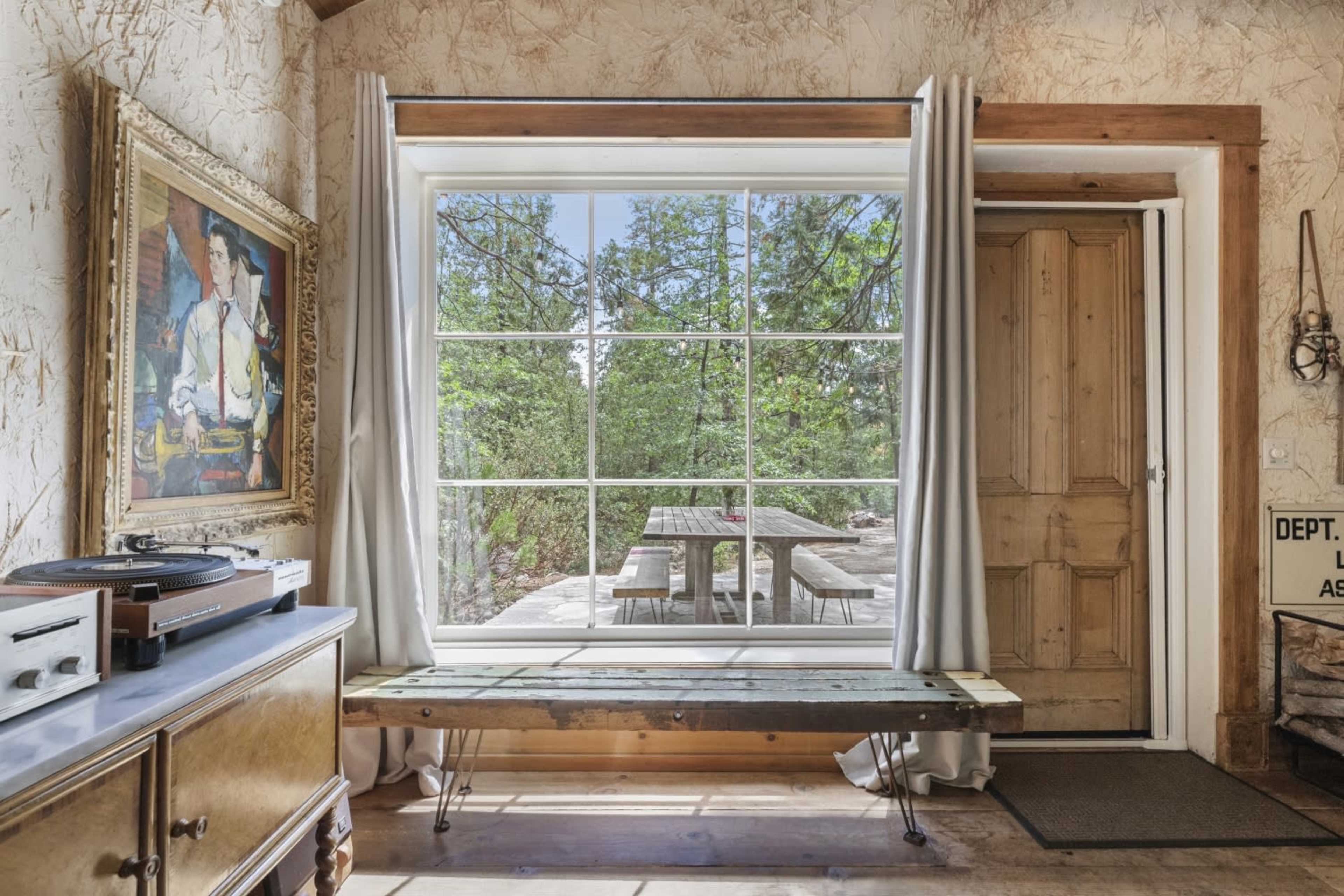 A large window framed by curtains overlooks a wooden table on a patio surrounded by trees.
