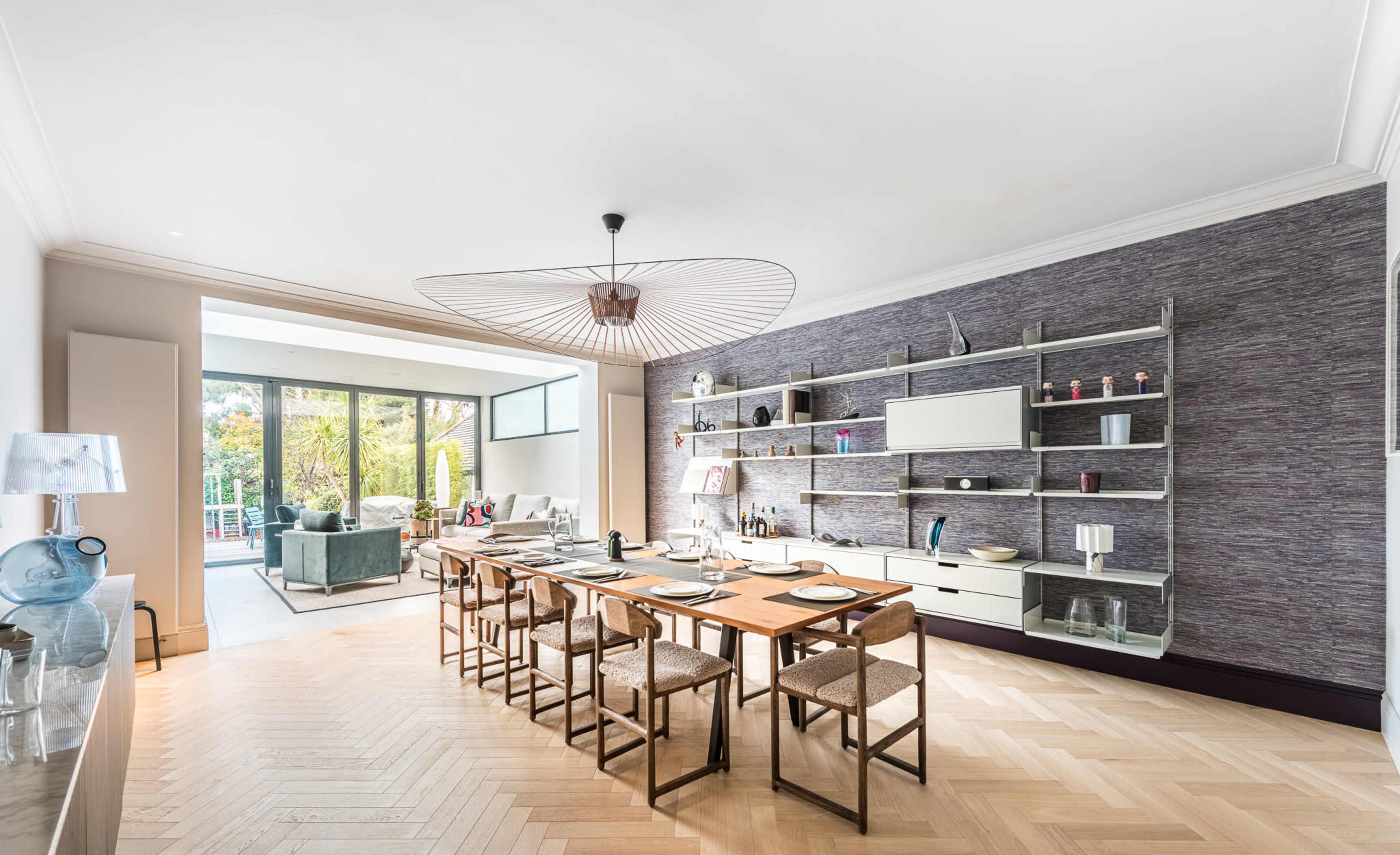 A modern dining area features a long wooden table with woven chairs, and shelves displaying decorative items, leading to a bright living space with large windows in the background.