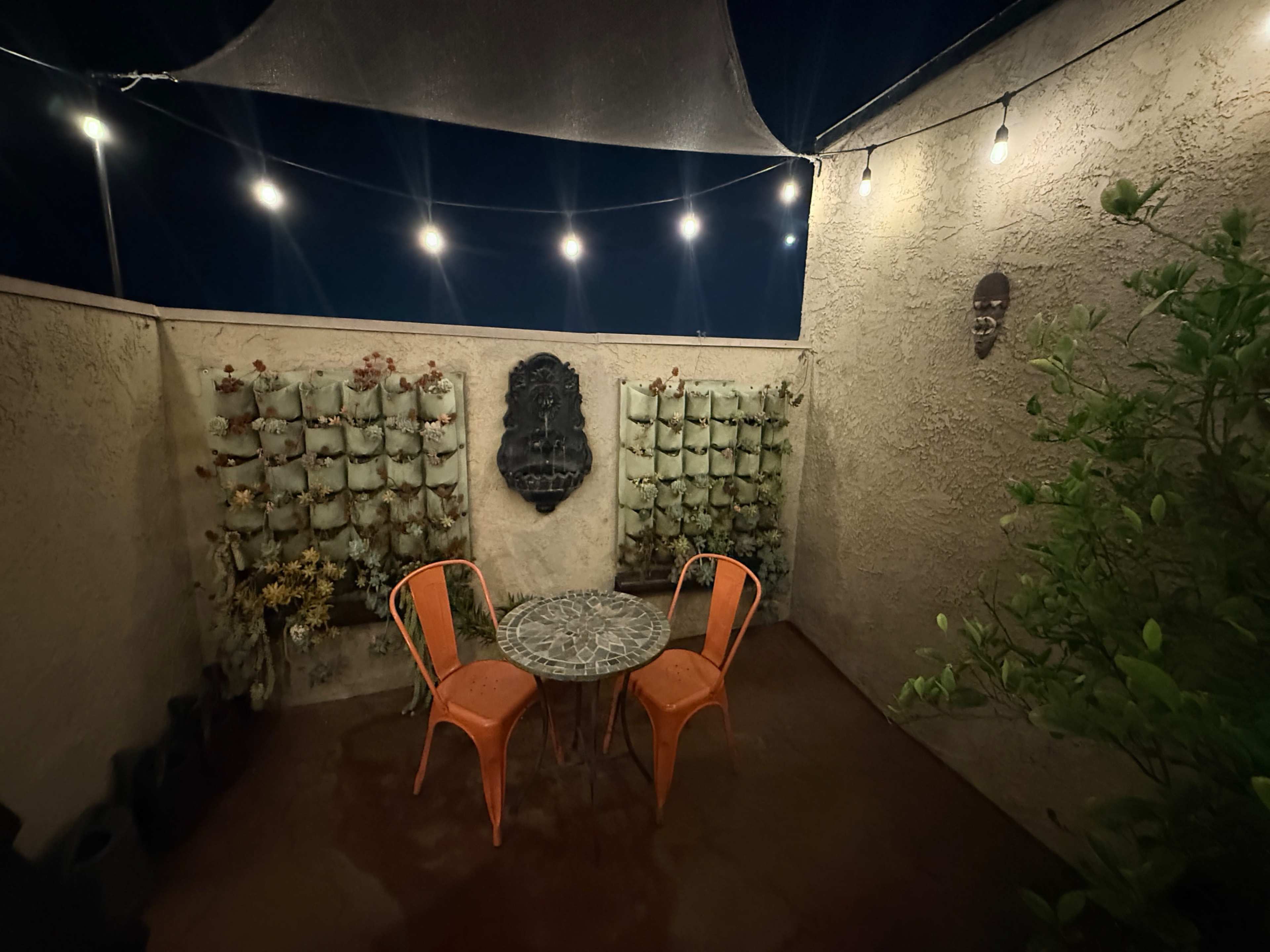 The image shows a small outdoor patio with two orange chairs and a round table, surrounded by planters on the wall and overhead string lights illuminated in the nighttime.