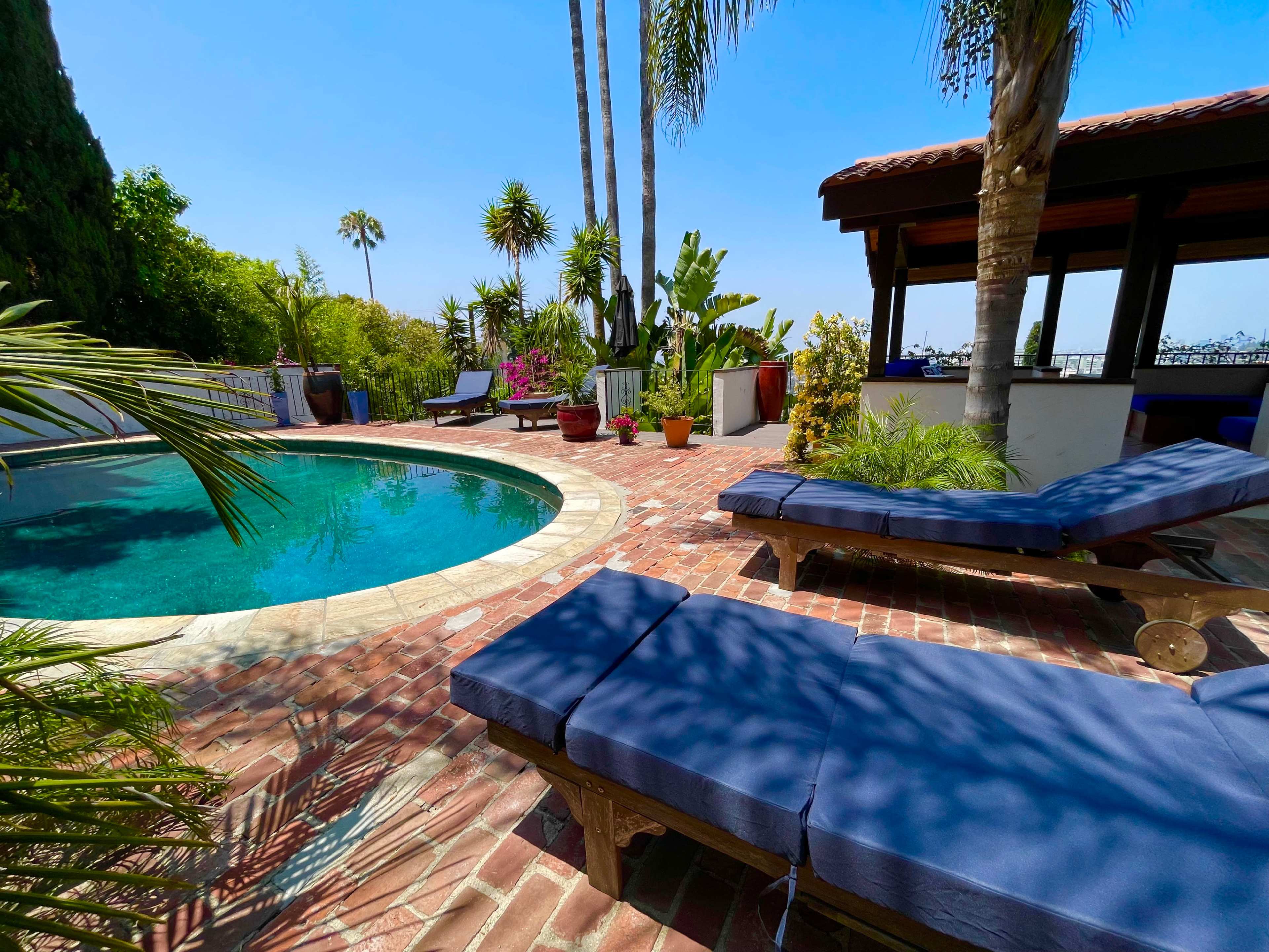 A swimming pool is surrounded by brick paving, lounge chairs, and various tropical plants in a sunny outdoor setting.