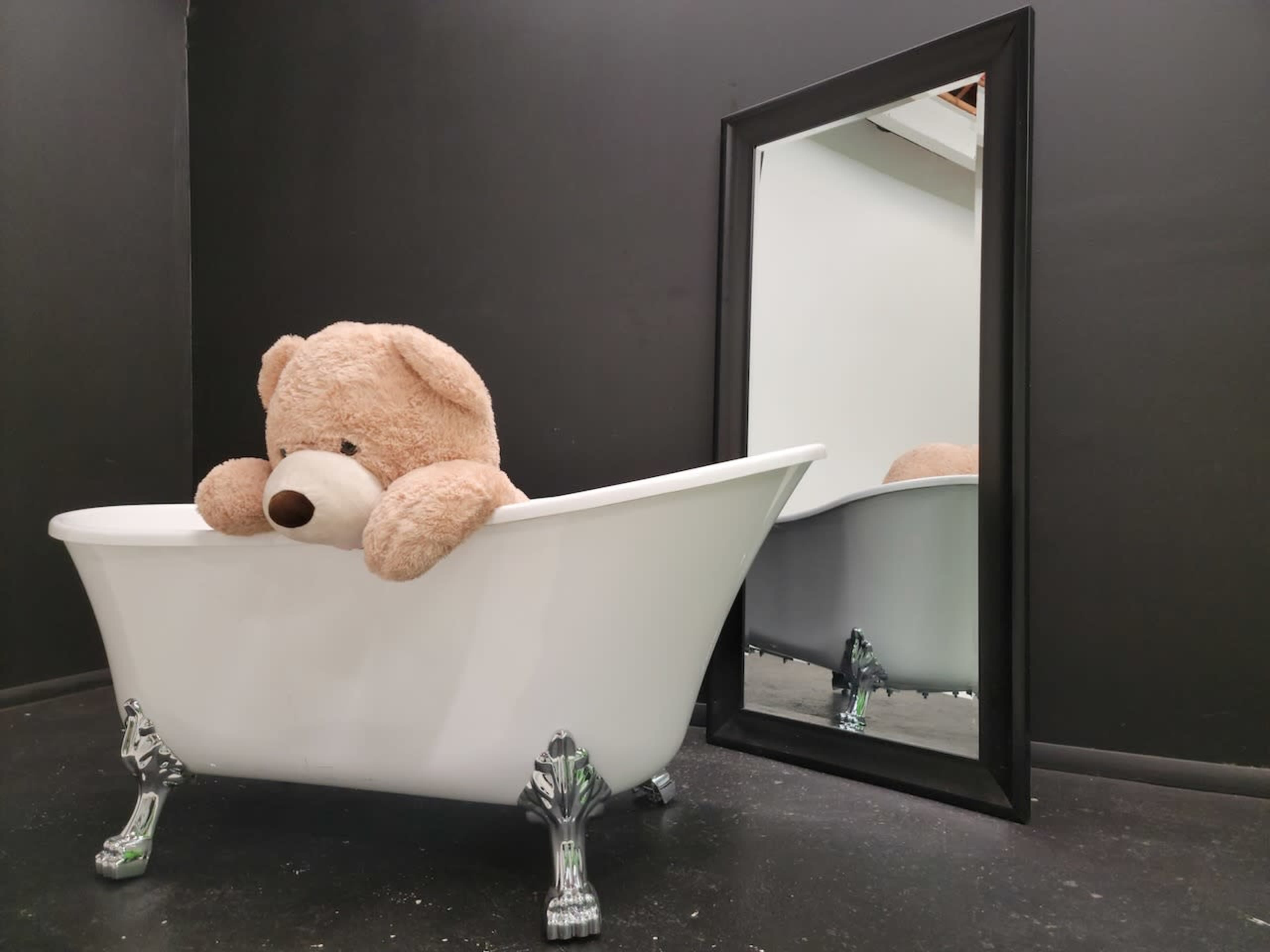A large teddy bear rests in a white freestanding bathtub next to a black-framed mirror reflecting the scene.