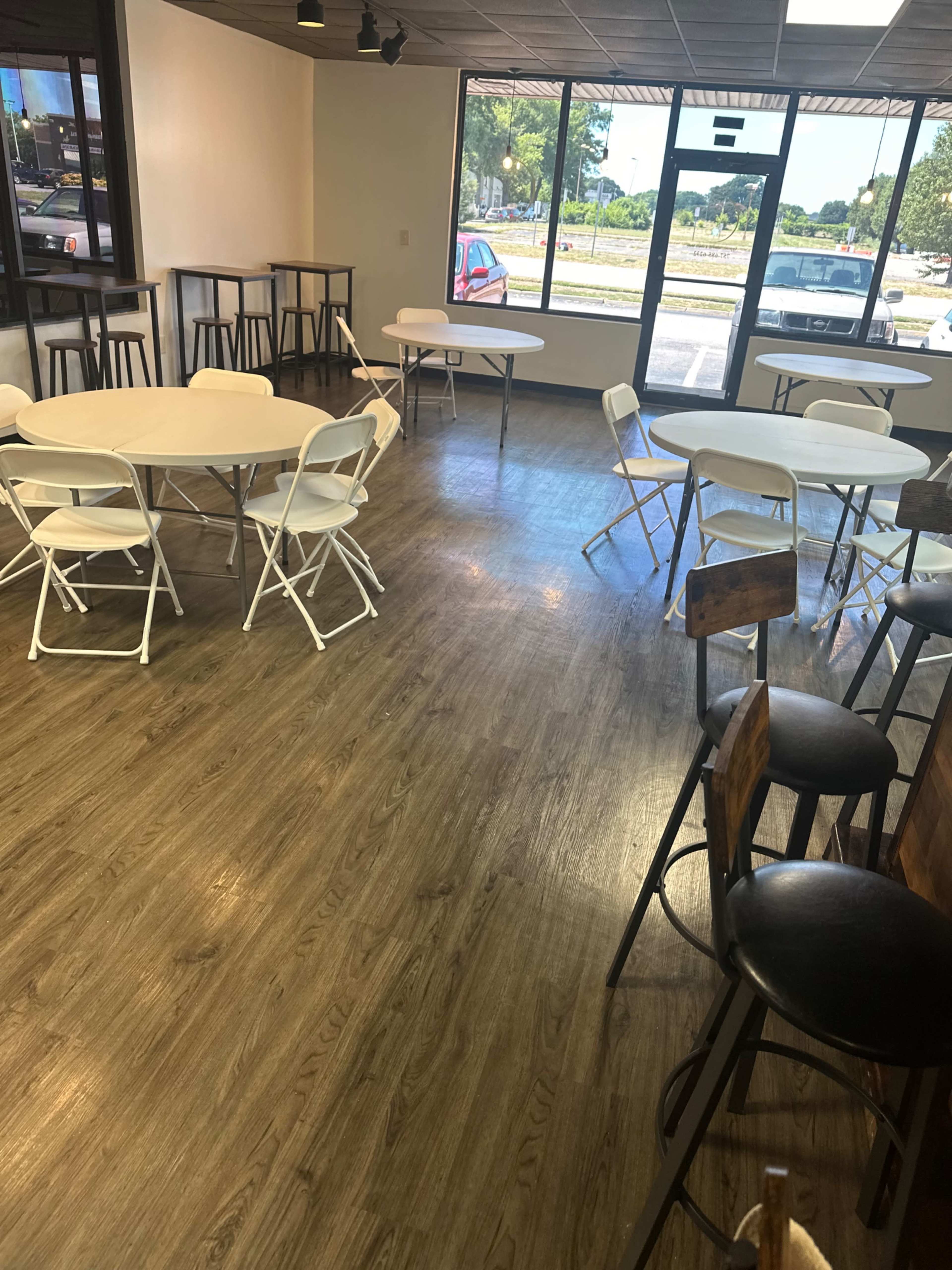 A spacious room with several round and rectangular tables and chairs arranged on a wooden floor, with large windows letting in natural light.