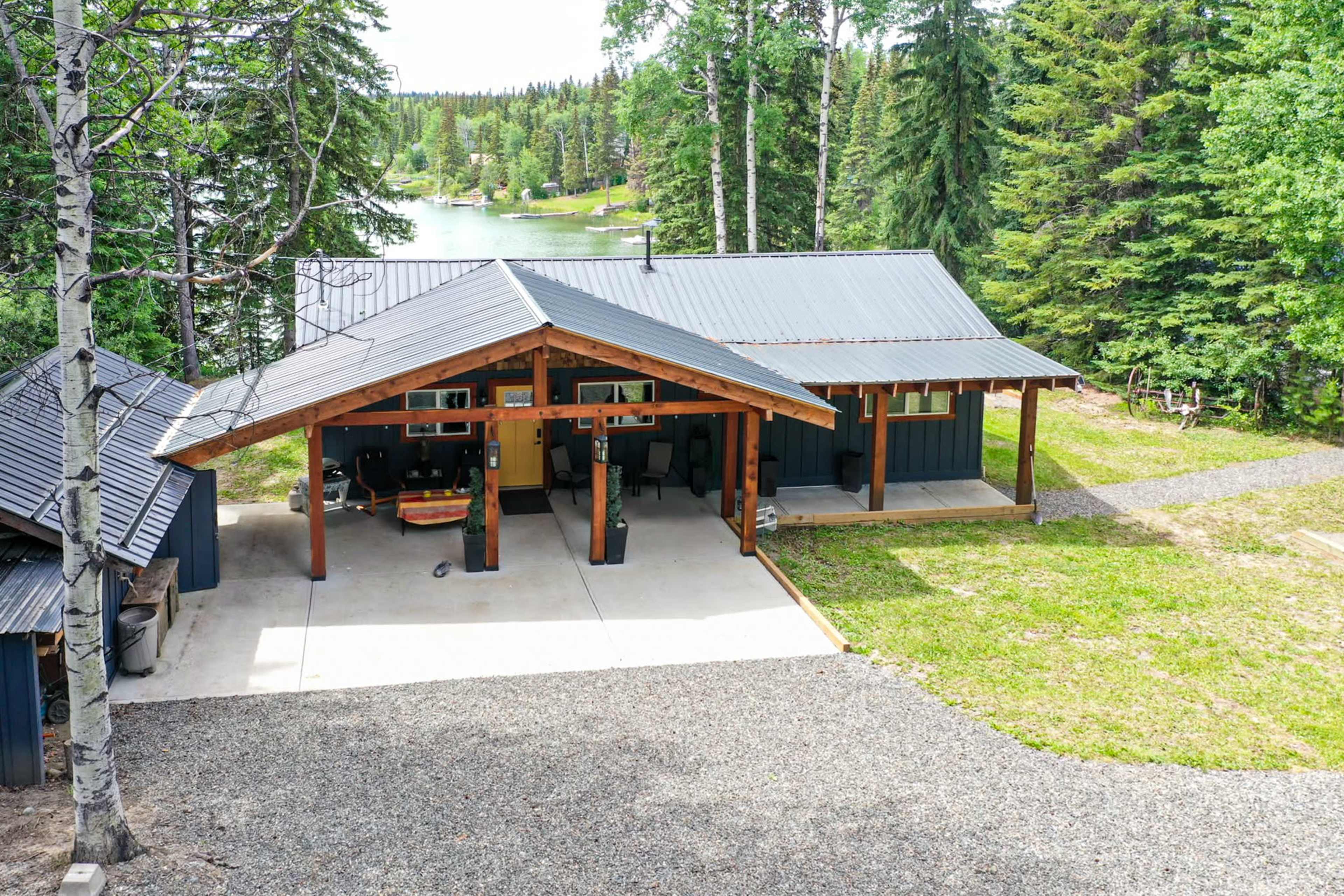 A wooden cabin with a metal roof is situated near a lake, surrounded by tall trees and a gravel pathway.