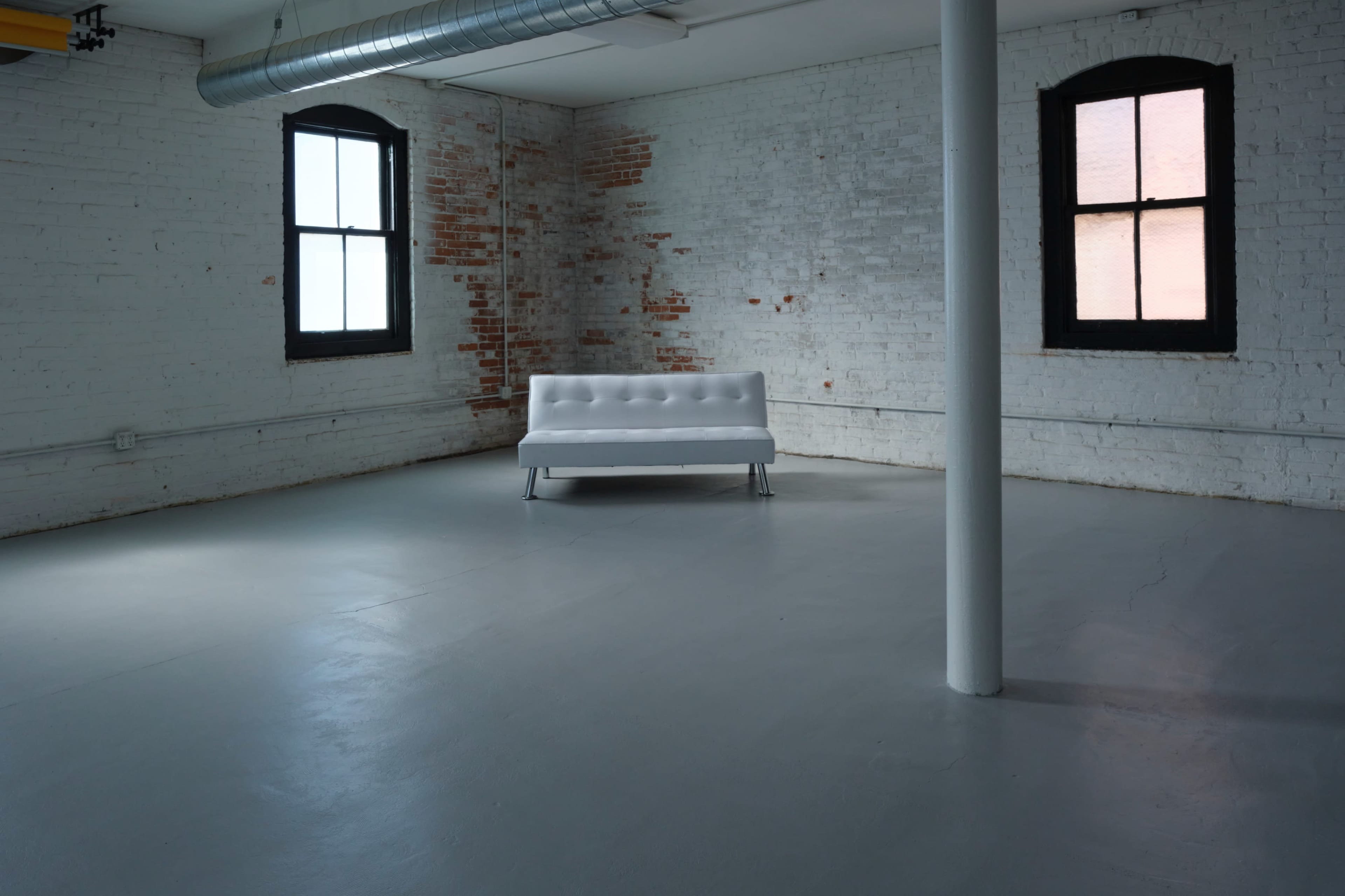 A minimalist room with white brick walls, two windows, a concrete floor, and a white sofa positioned in the center.