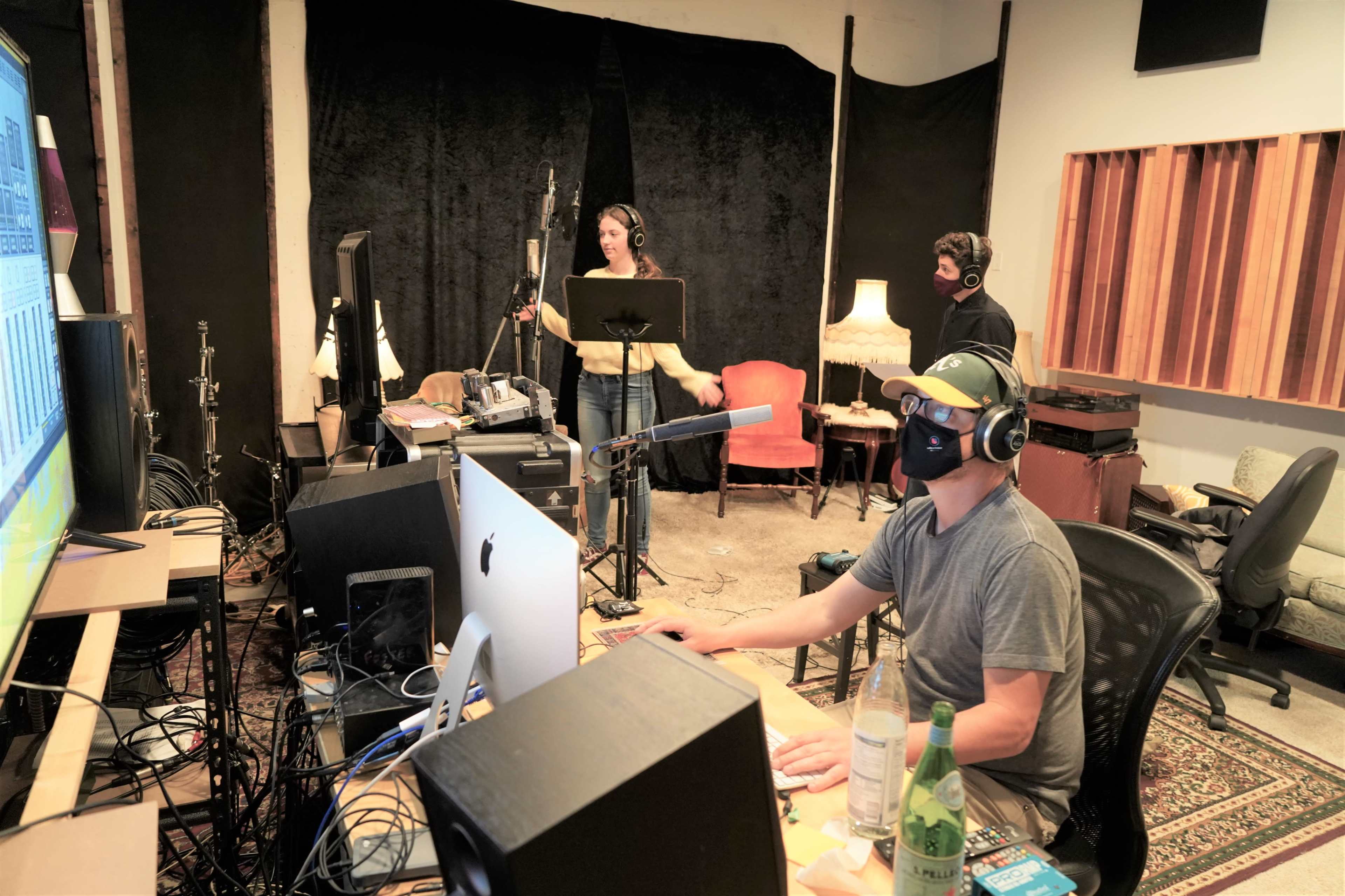A seated person works at a computer while two others record vocals in a sound studio.