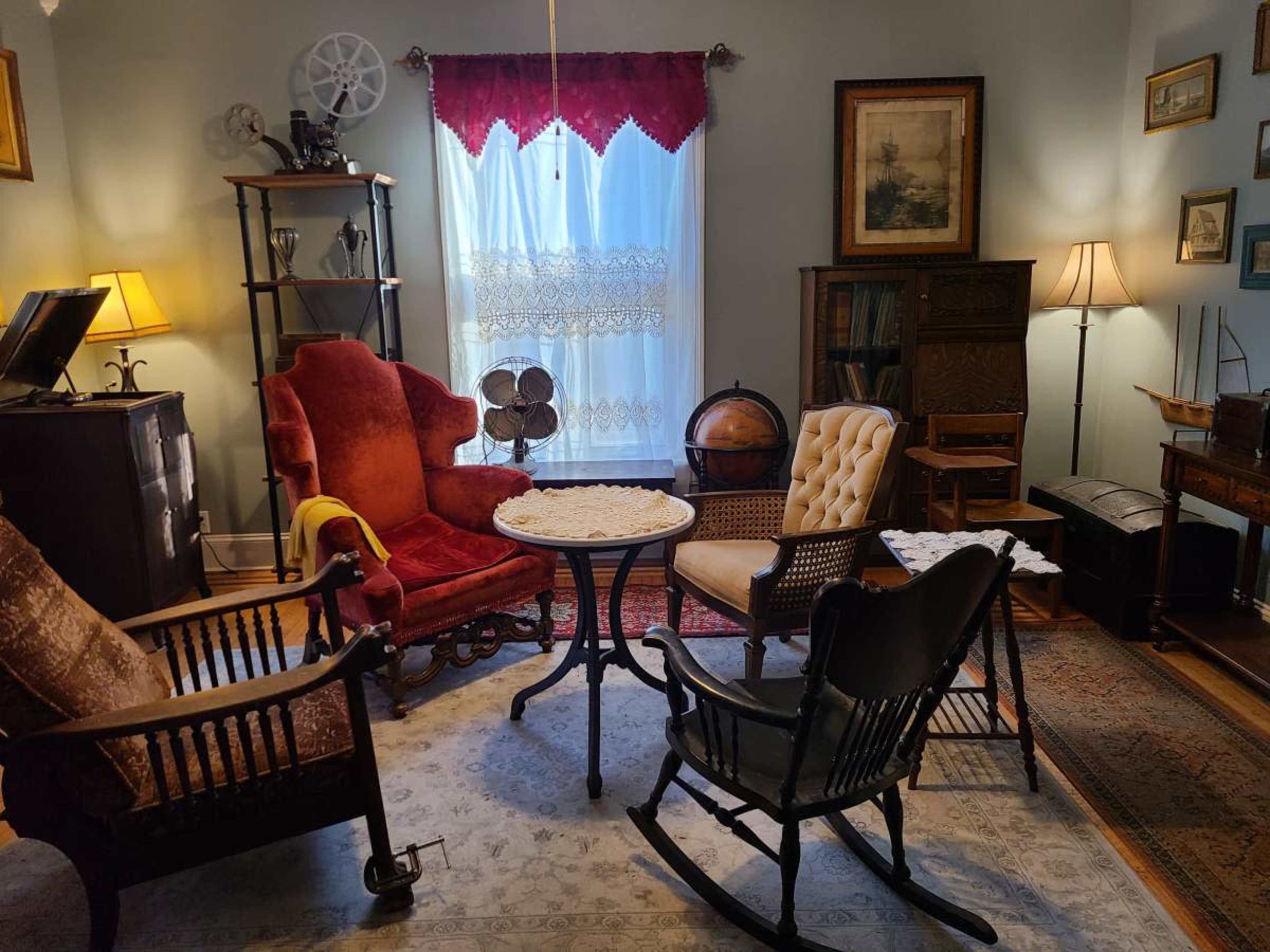 The room features an arrangement of vintage furniture, including rocking chairs and a round table, illuminated by soft lamp light and natural light from a window.