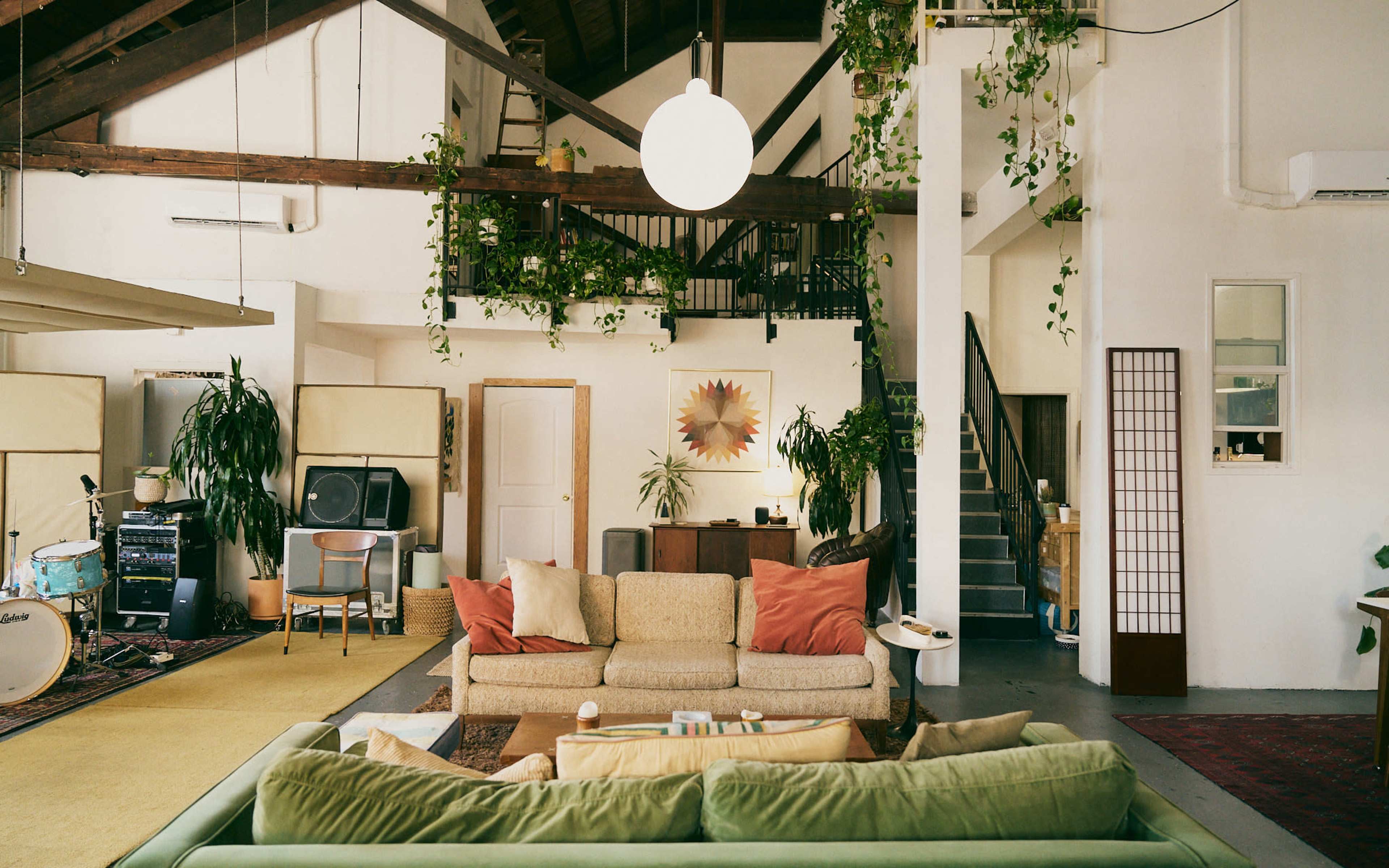 The image shows a spacious, modern living area featuring two sofas, plants, a staircase, and various furnishings in a bright, open layout.