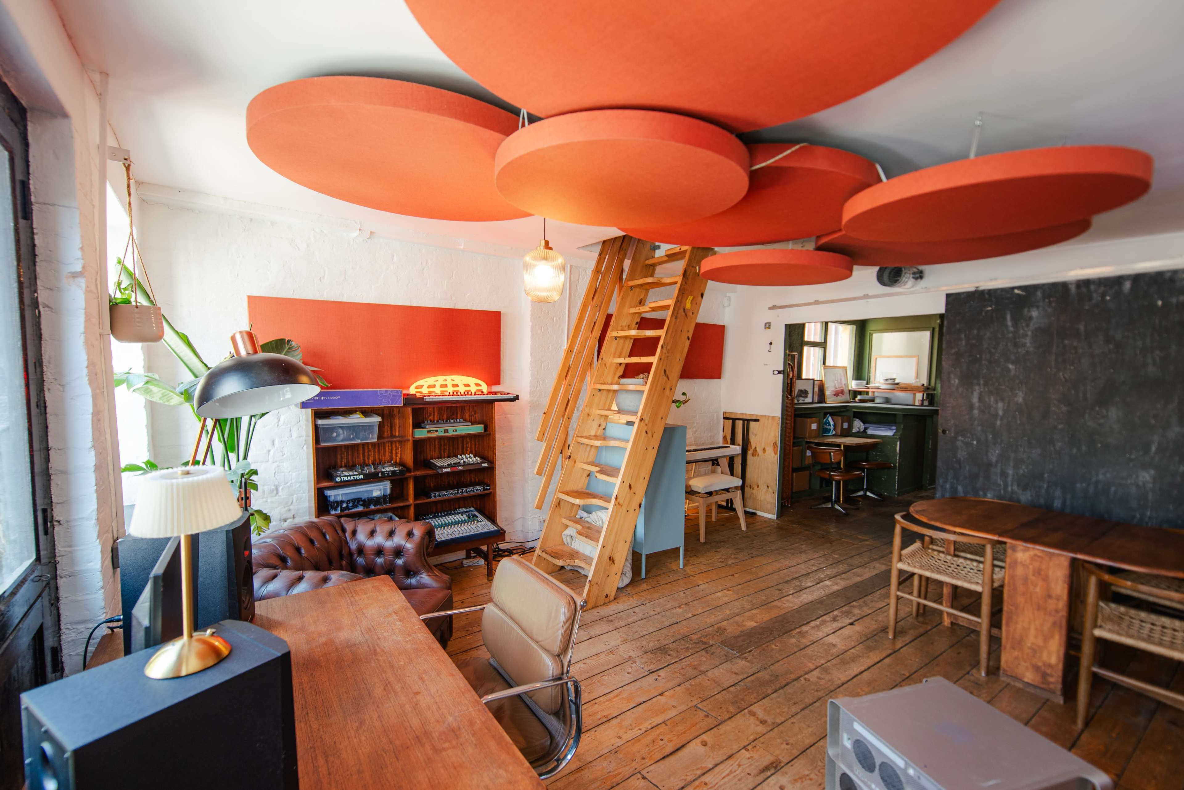 The image shows a bright, open room with wooden floors, a staircase leading to a loft, and colorful circular acoustic panels hanging from the ceiling.