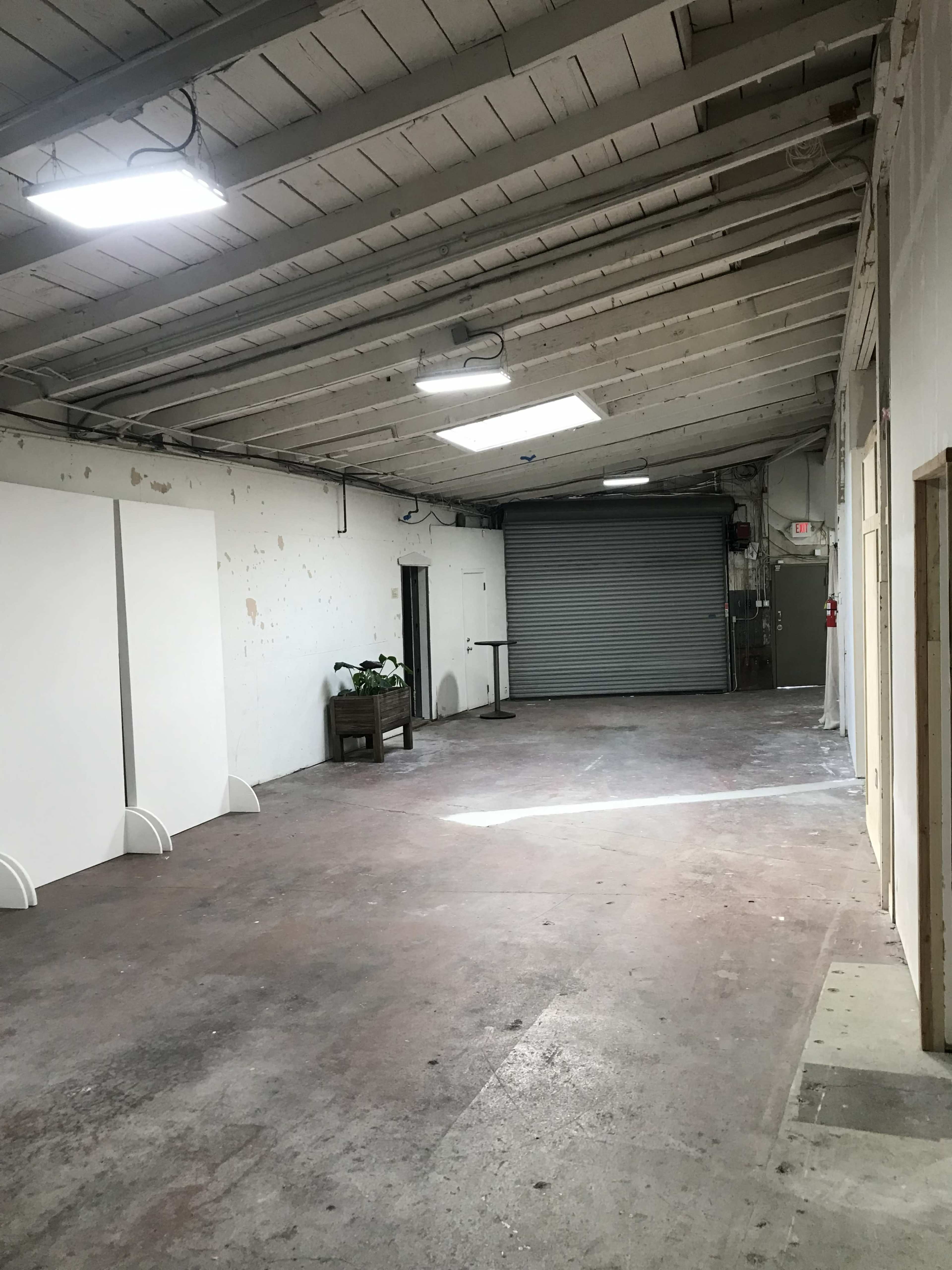 The image shows a spacious, industrial-style room with a high ceiling, featuring concrete floors, a large roll-up door, and minimal furnishings.