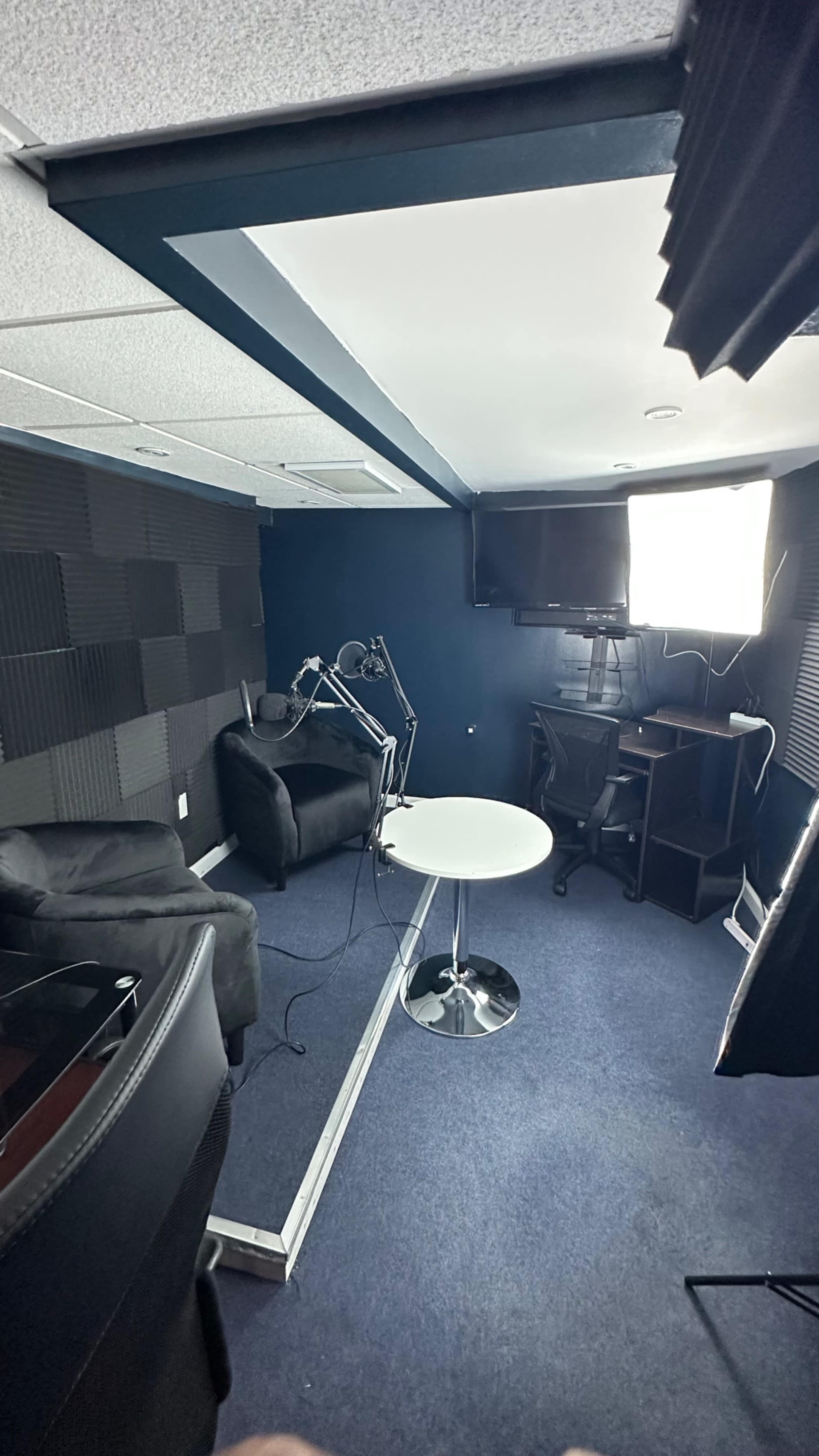 A small, dimly lit recording studio with soundproof walls, a table in the center, and black furniture arranged around it.