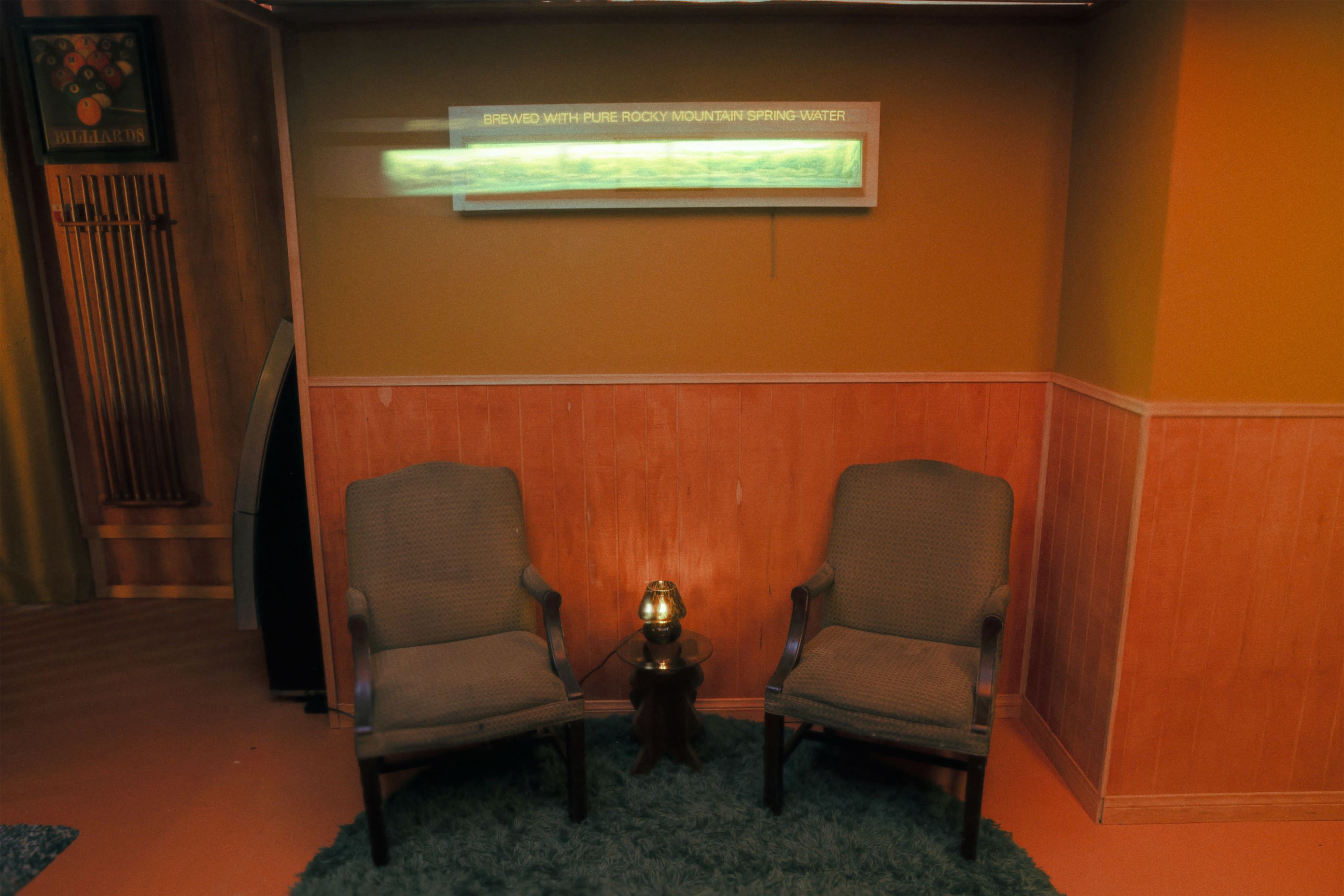Two upholstered chairs are positioned next to a small table with a lamp, set against a backdrop of wooden paneling and a wall-mounted sign.