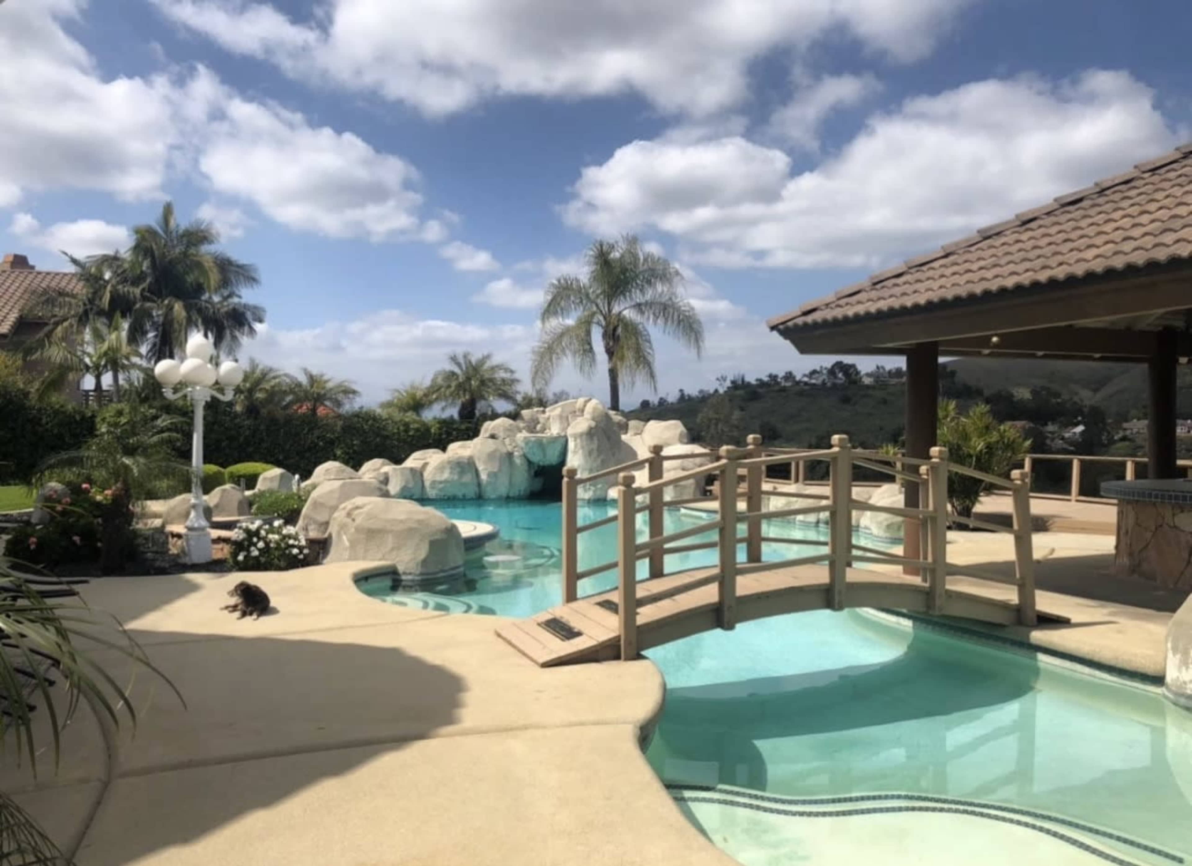 The image shows a swimming pool area featuring a rock formation, a bridge, and landscaped greenery under a partly cloudy sky.