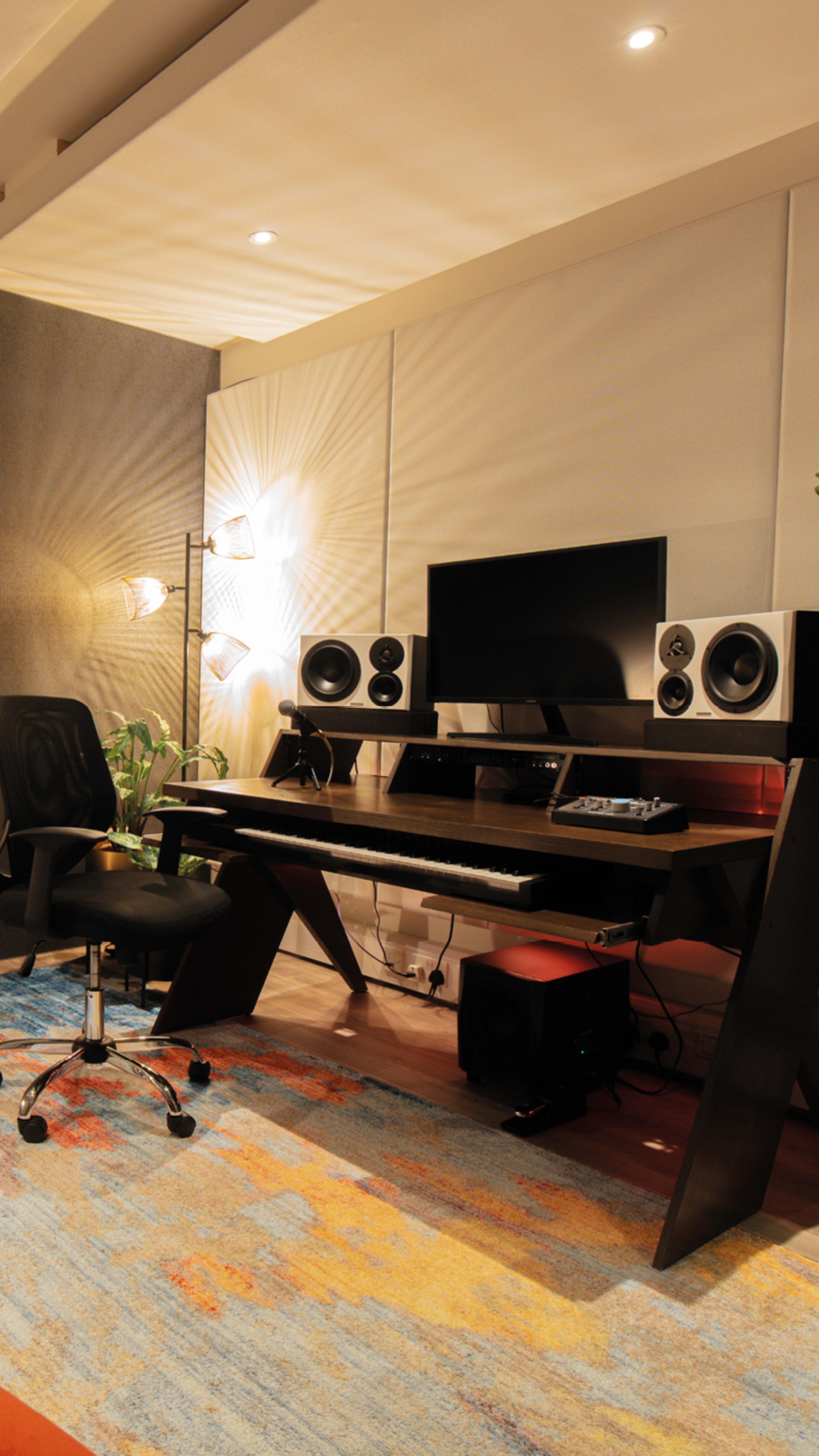 The image shows a modern home studio setup featuring a keyboard, monitors, and a computer display on a sleek desk, accompanied by a swivel chair and decorative lighting.