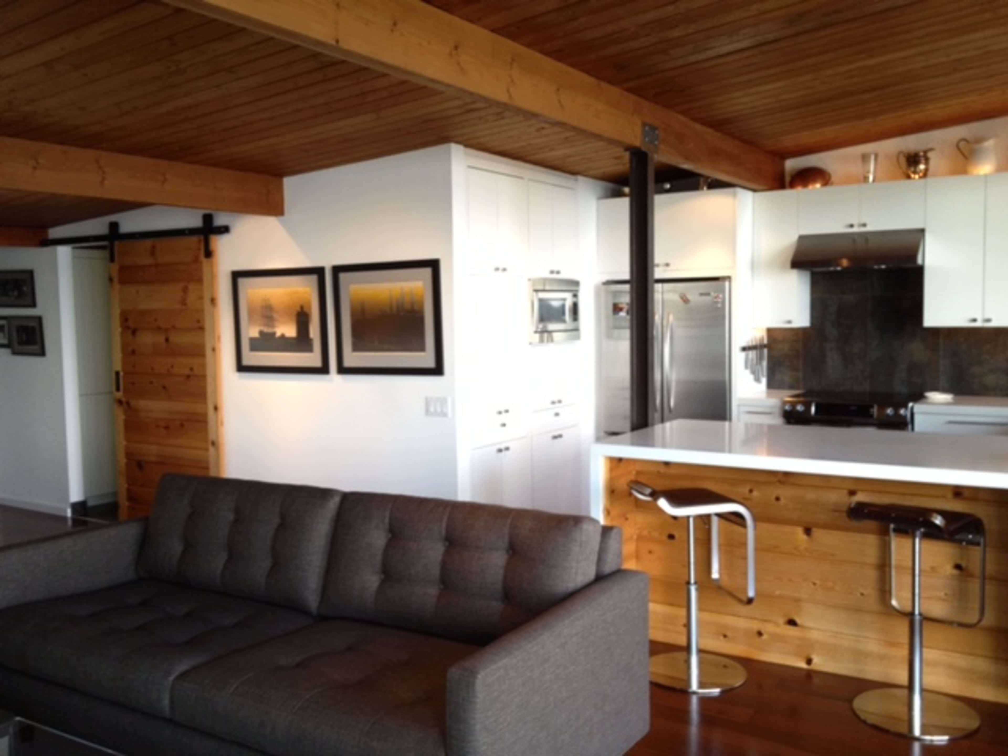 A modern open-plan living area with a gray sofa, wooden ceiling beams, and a kitchen featuring white cabinetry and stainless steel appliances.