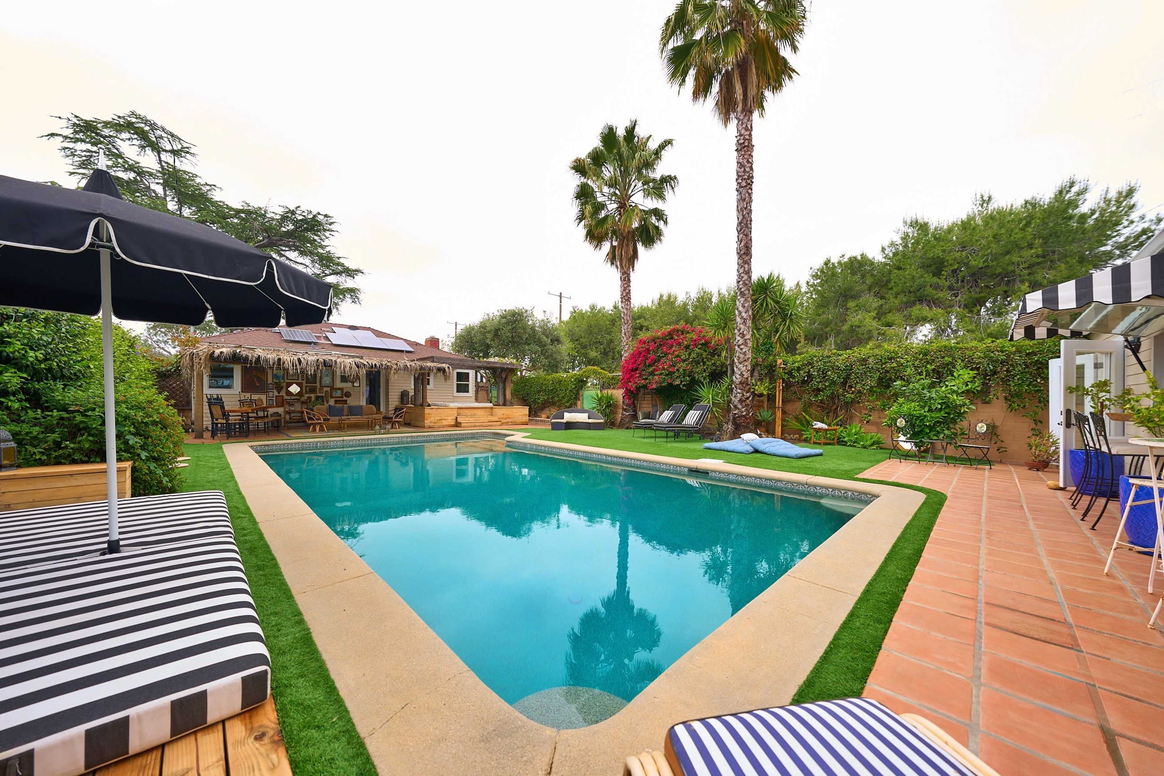 A rectangular swimming pool is surrounded by lounge chairs, palm trees, and lush greenery in a backyard setting.