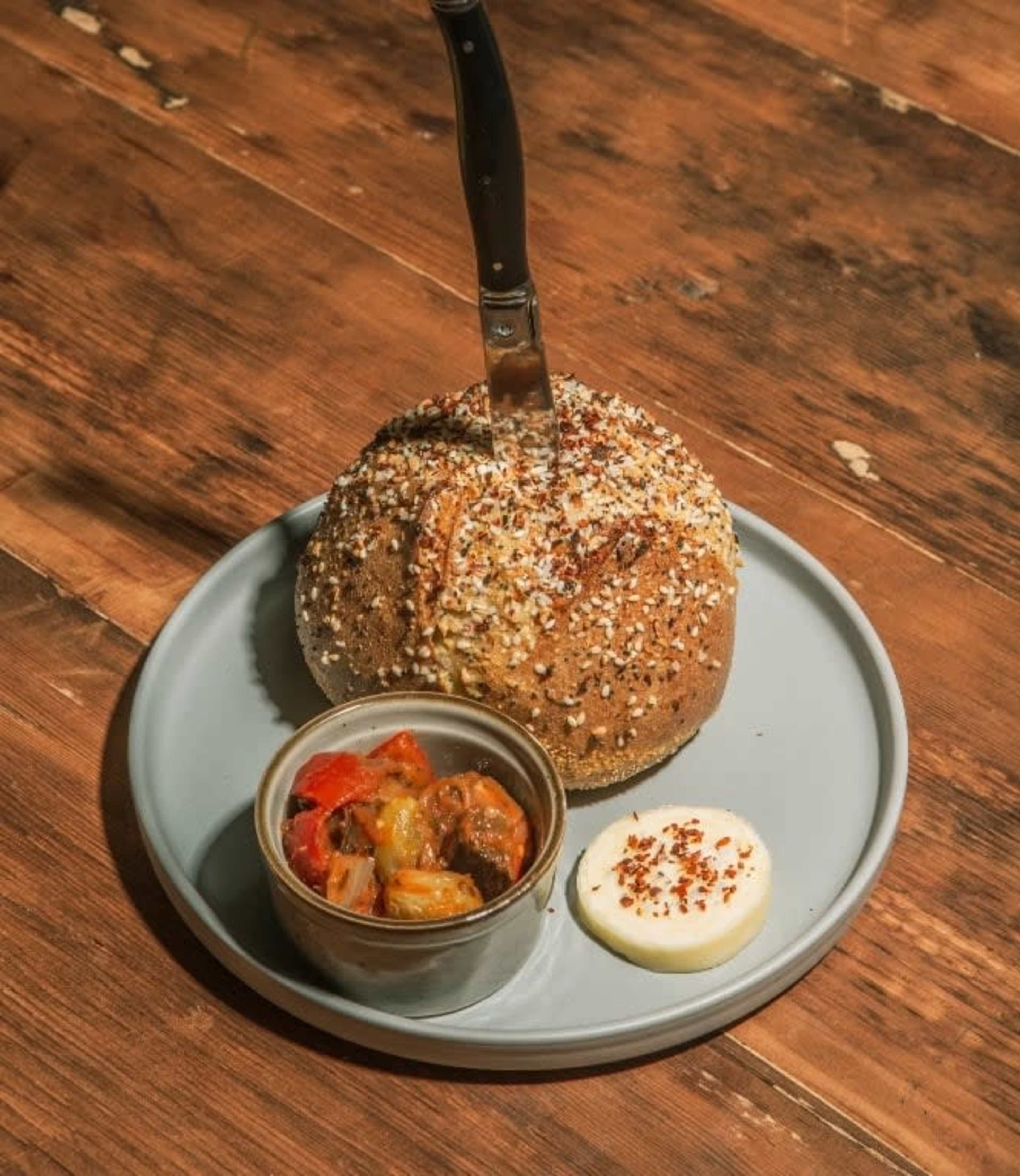 A round loaf of bread topped with sesame seeds is served on a gray plate alongside a small bowl of roasted vegetables and a dollop of creamy sauce, with a knife inserted into the bread.