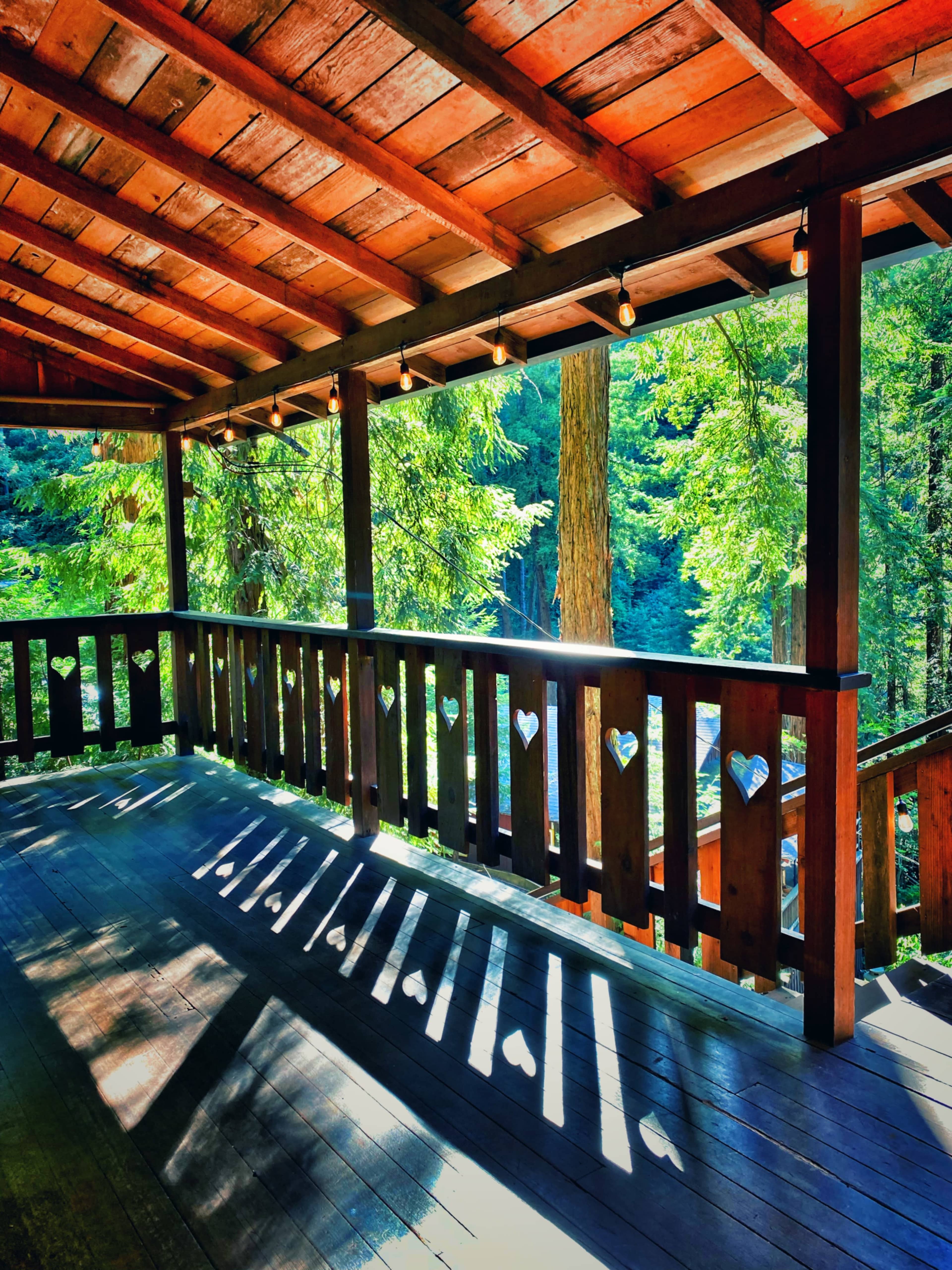 The image shows a wooden porch with heart-shaped cutouts in the railing, surrounded by lush greenery and dappled sunlight.