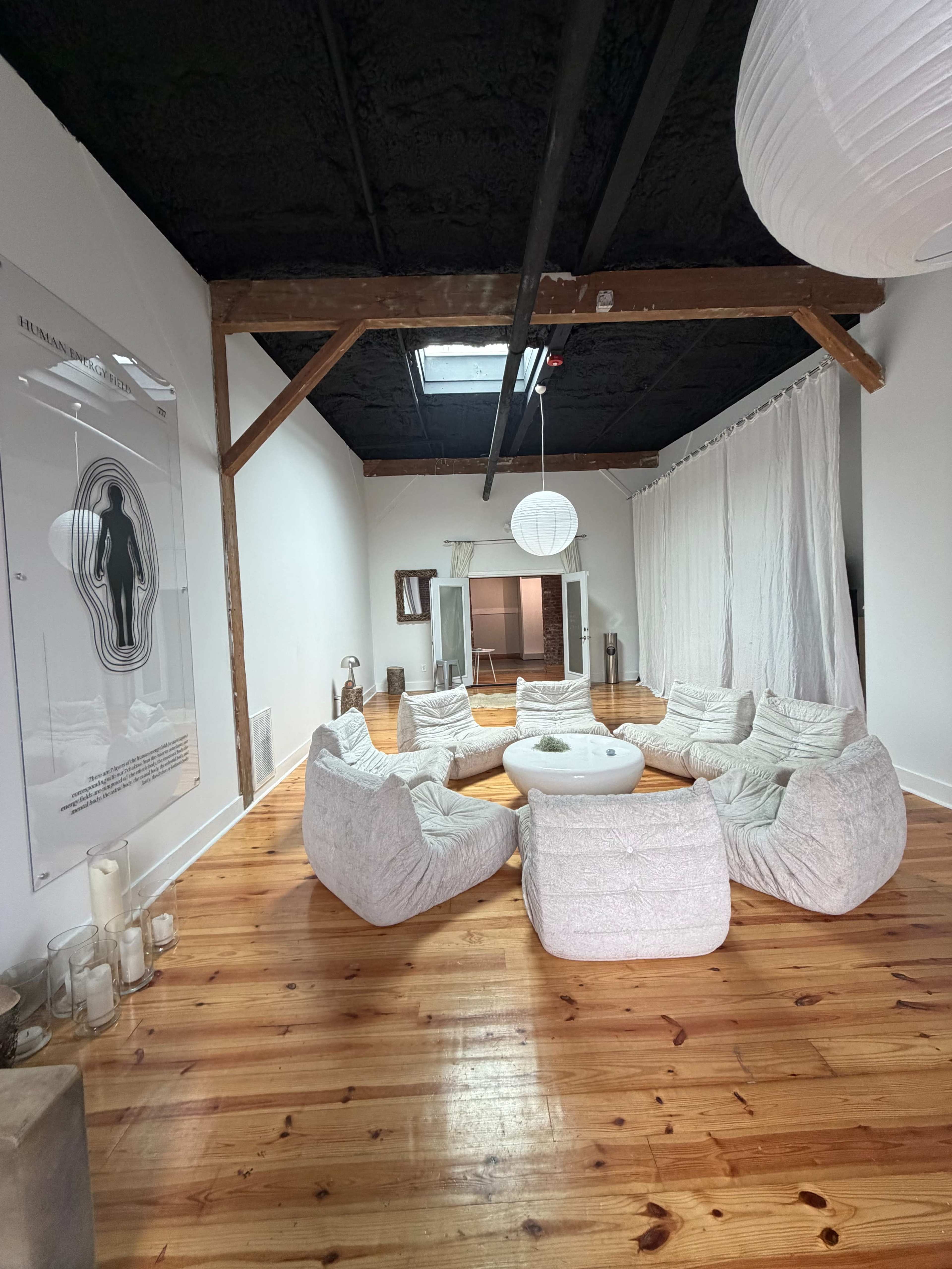 A spacious room with wooden floors features a circular arrangement of soft chairs around a central table, complemented by hanging light fixtures and white drapes.