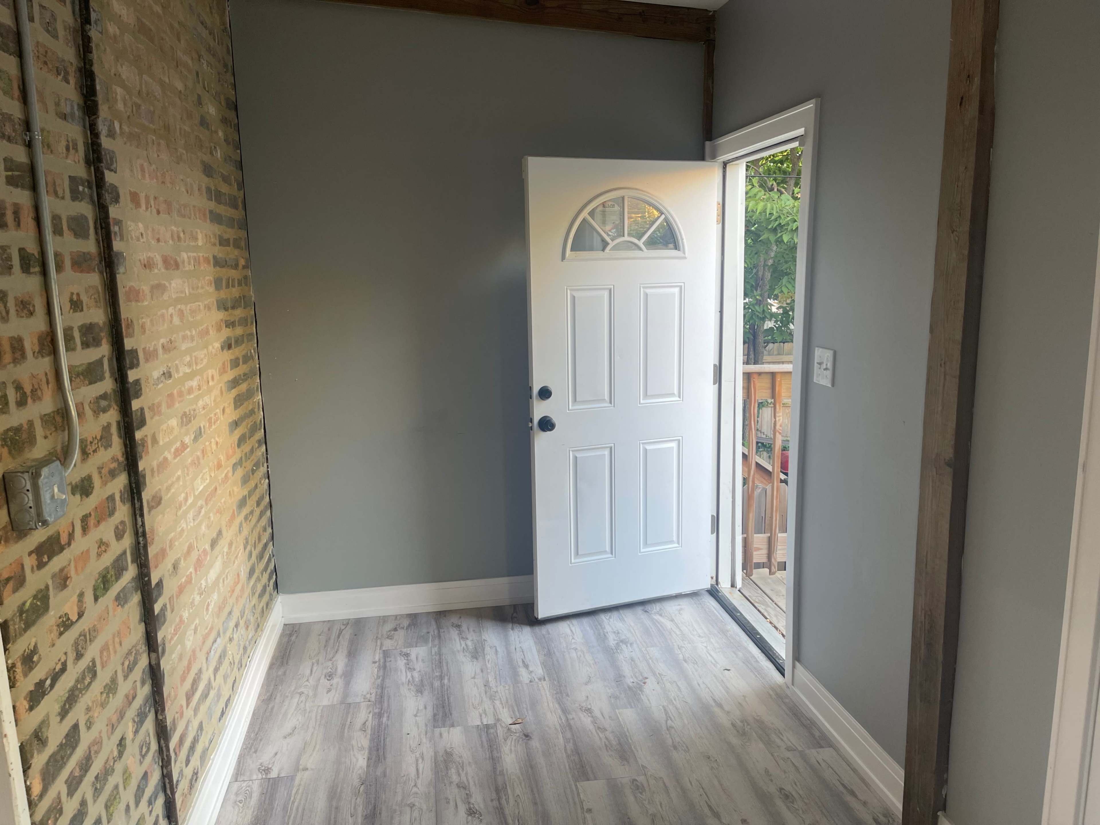 A room with a brick accent wall and an open white door leading to an outdoor deck.