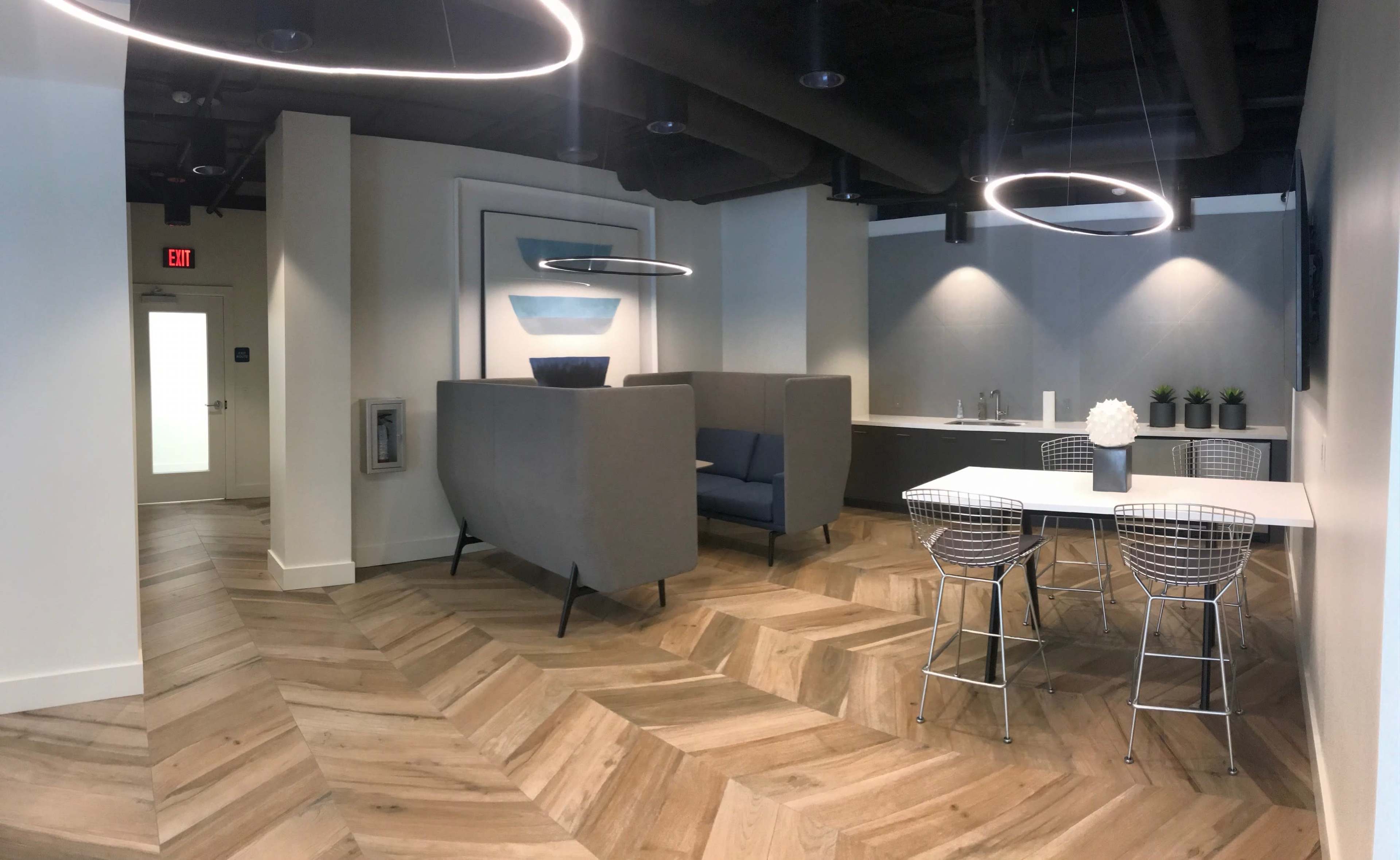 A modern office space featuring a lounge area with a gray sofa, a kitchenette, and a dining table surrounded by wire chairs, all set on a chevron-patterned wooden floor.