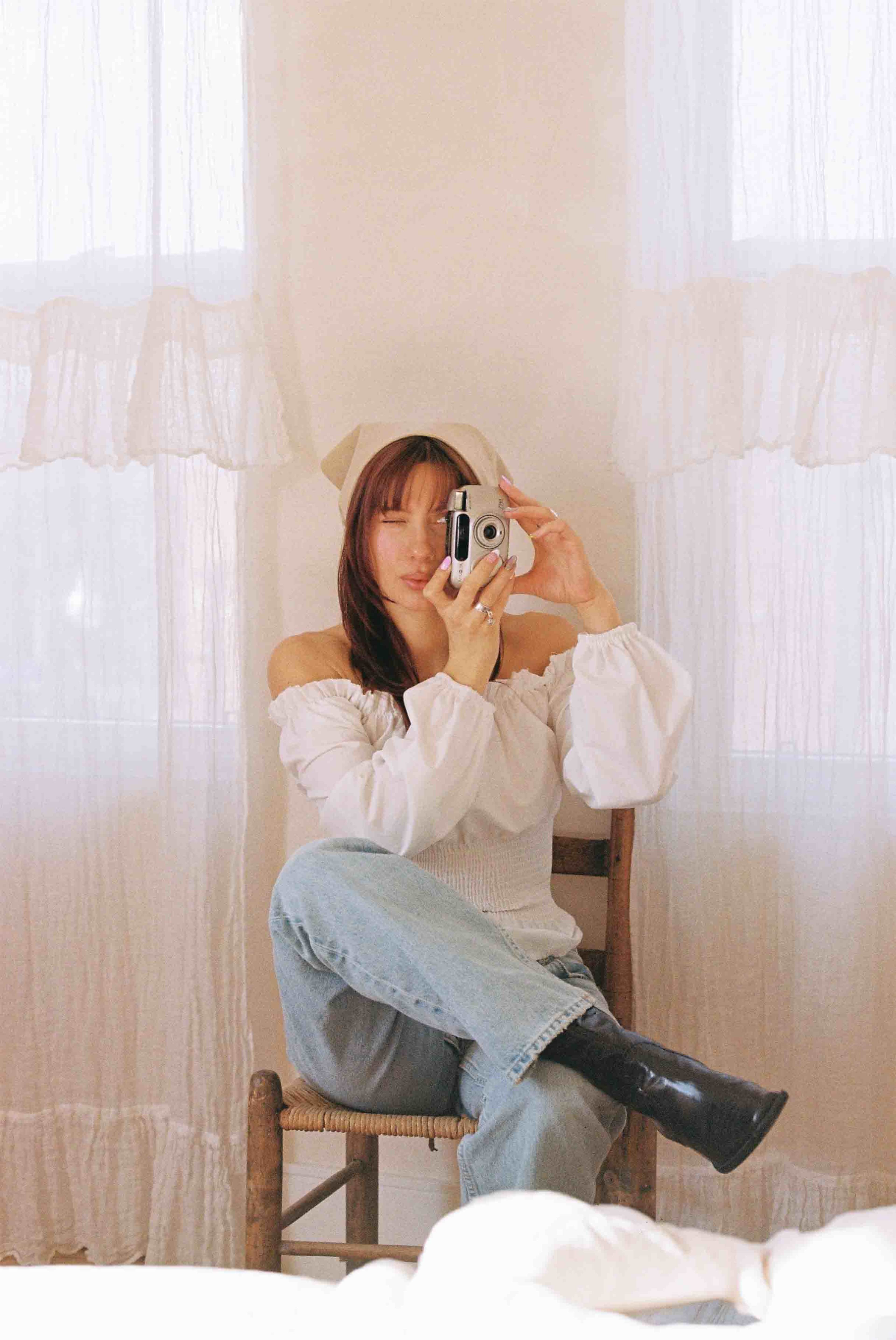 A person is sitting on a wooden chair, holding a camera and facing the viewer, in a room with light-colored curtains.