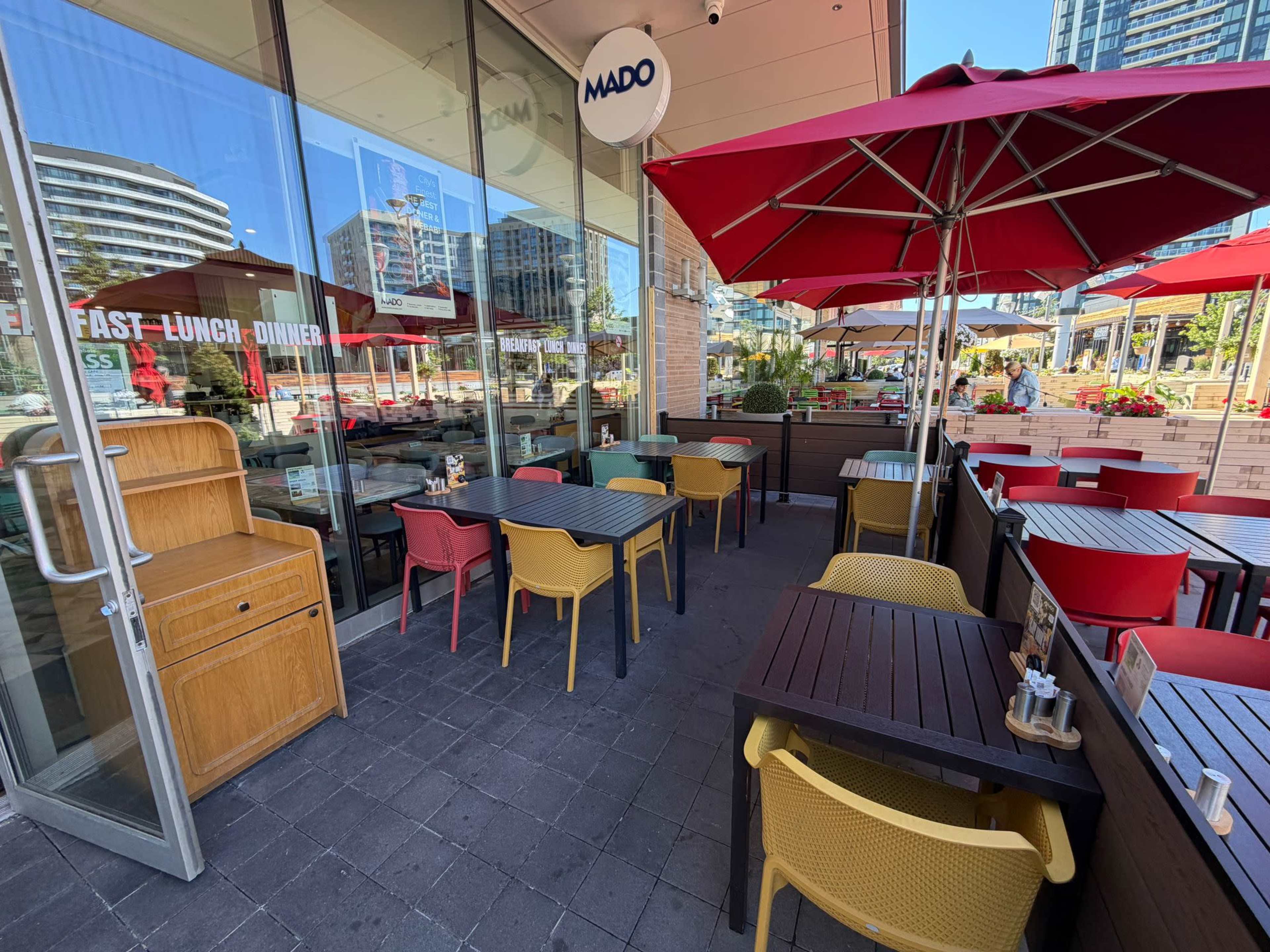 A restaurant patio features red and yellow chairs and umbrellas, with tables arranged along the exterior of a glass building.