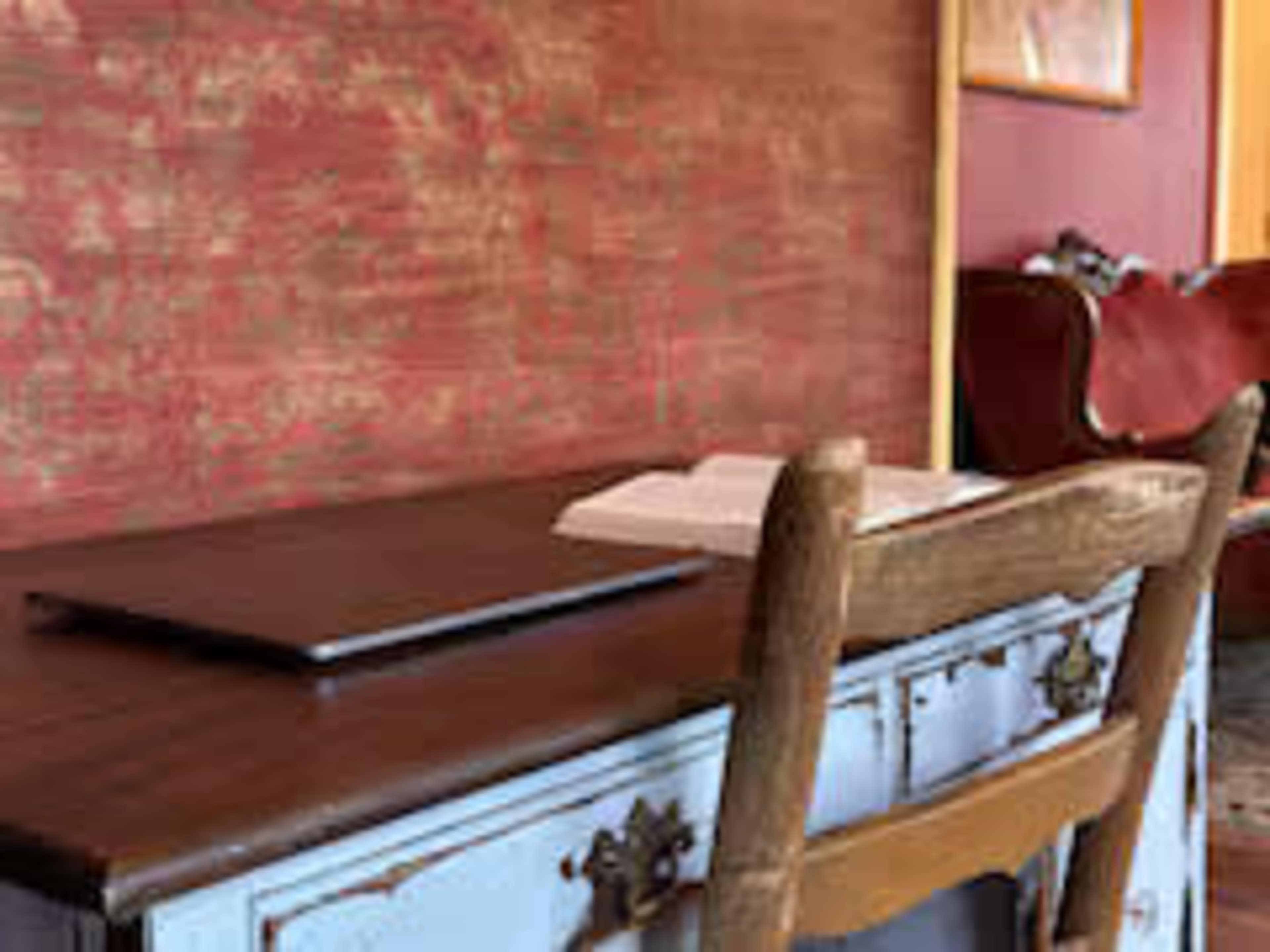 A wooden desk with a laptop and an open book sits against a textured red wall, with a chair positioned nearby.