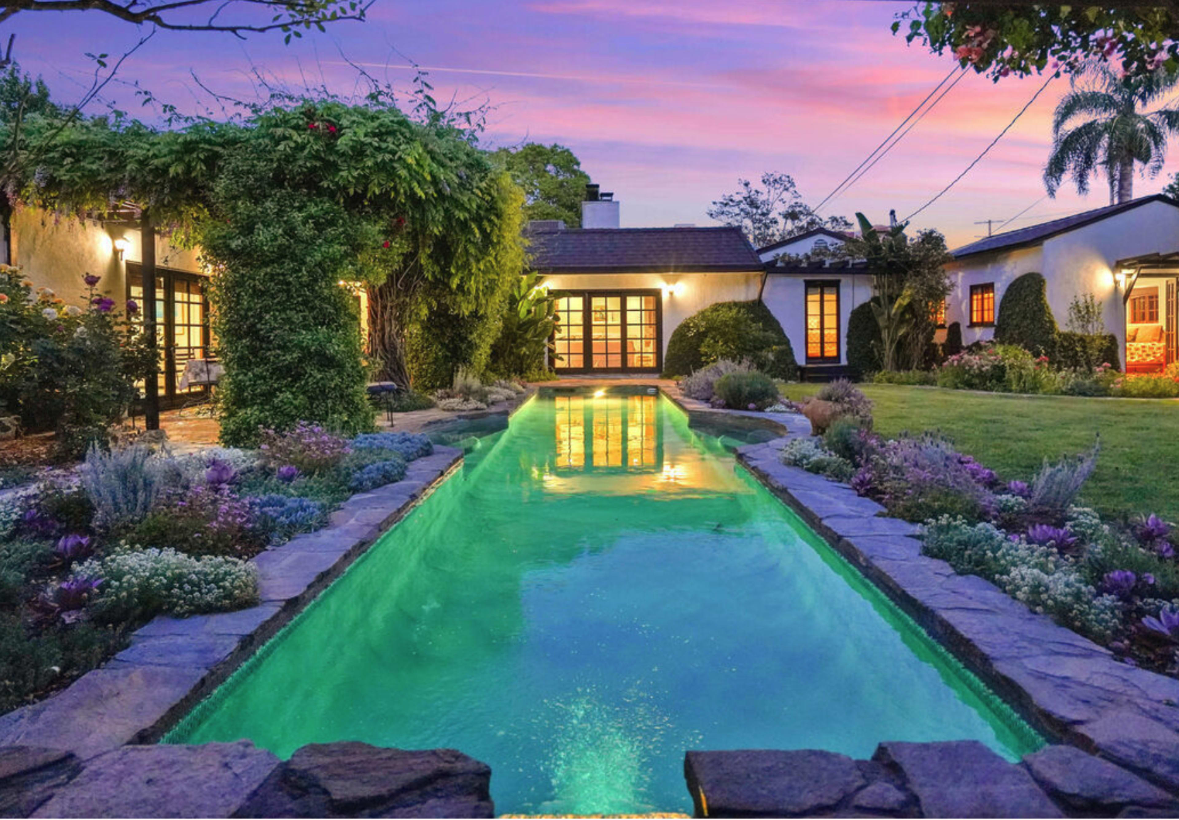 The image shows a tranquil backyard with a swimming pool reflecting the evening sky, bordered by landscaped gardens and a spacious home.