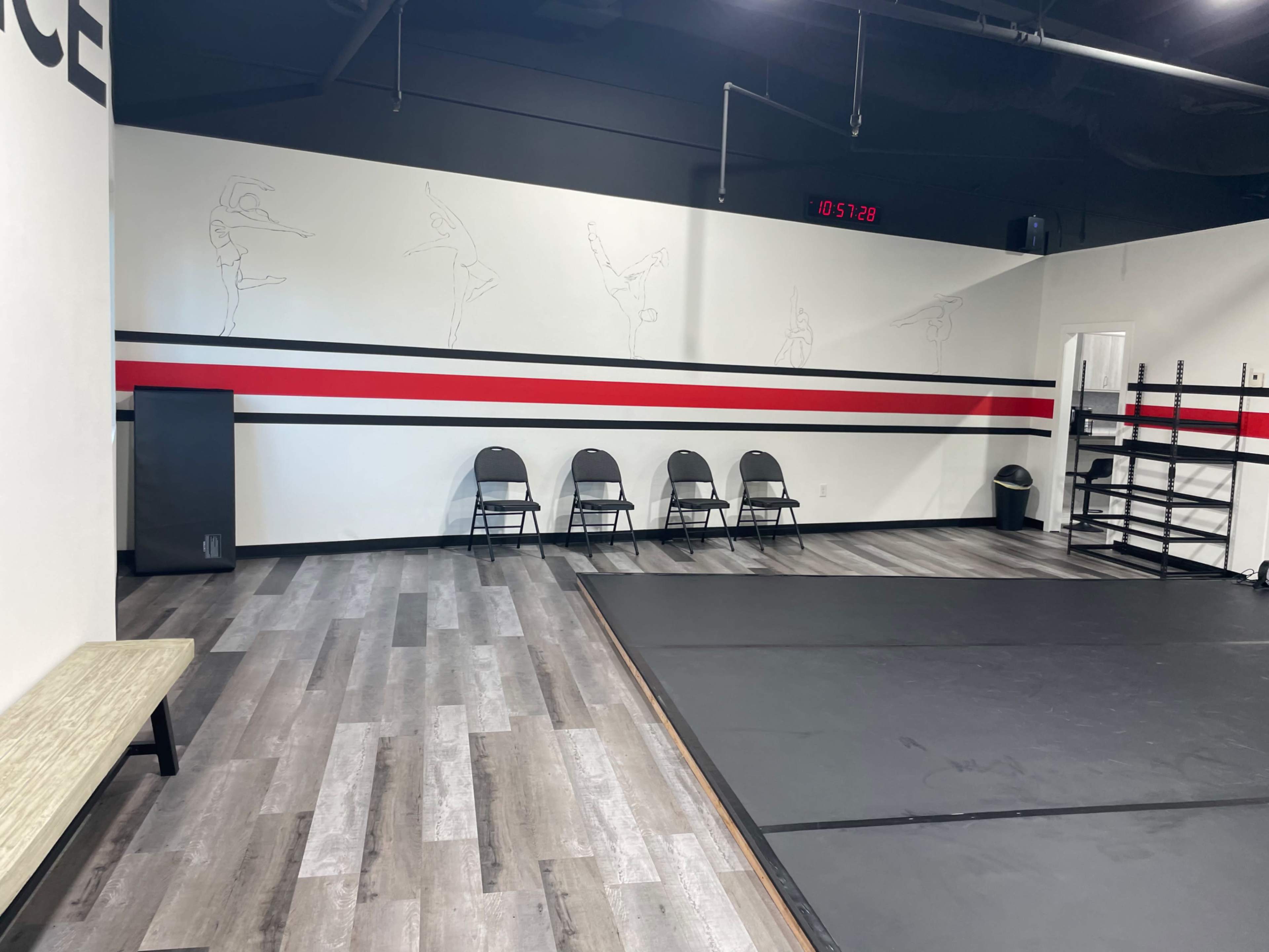 An empty dance studio with a mirrored wall, black mats on the floor, and several chairs arranged against the wall.