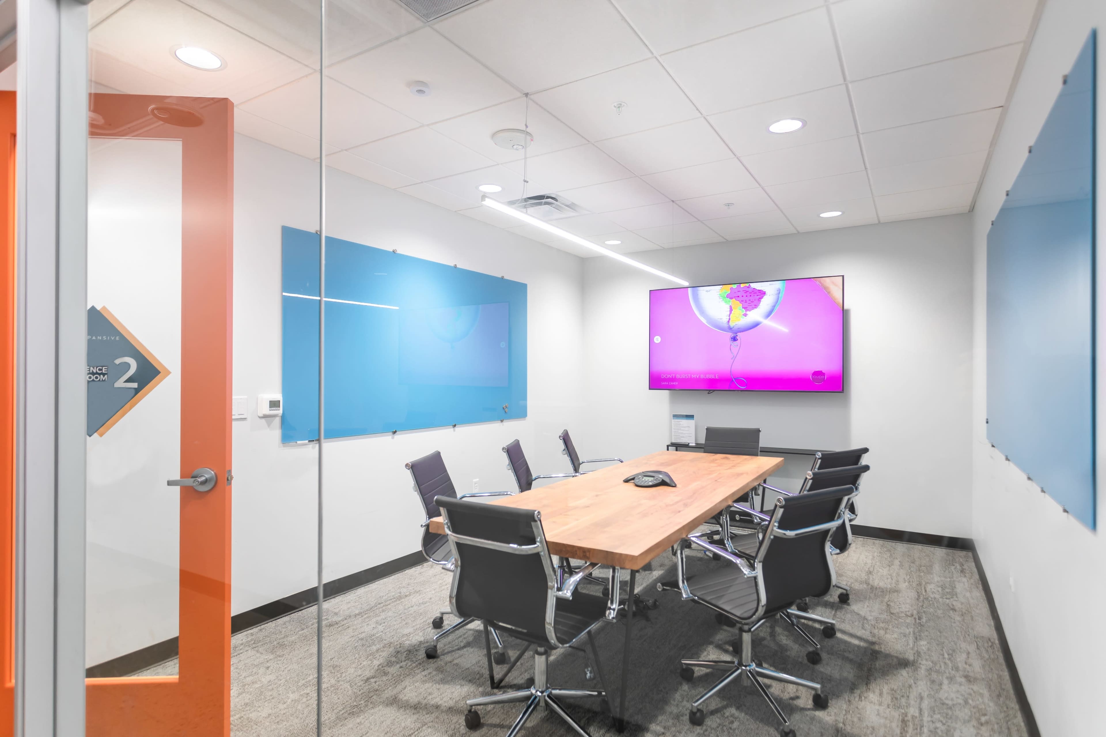 A modern conference room features a large wooden table surrounded by black chairs, with blue glass boards and a wall-mounted television displaying a colorful image.
