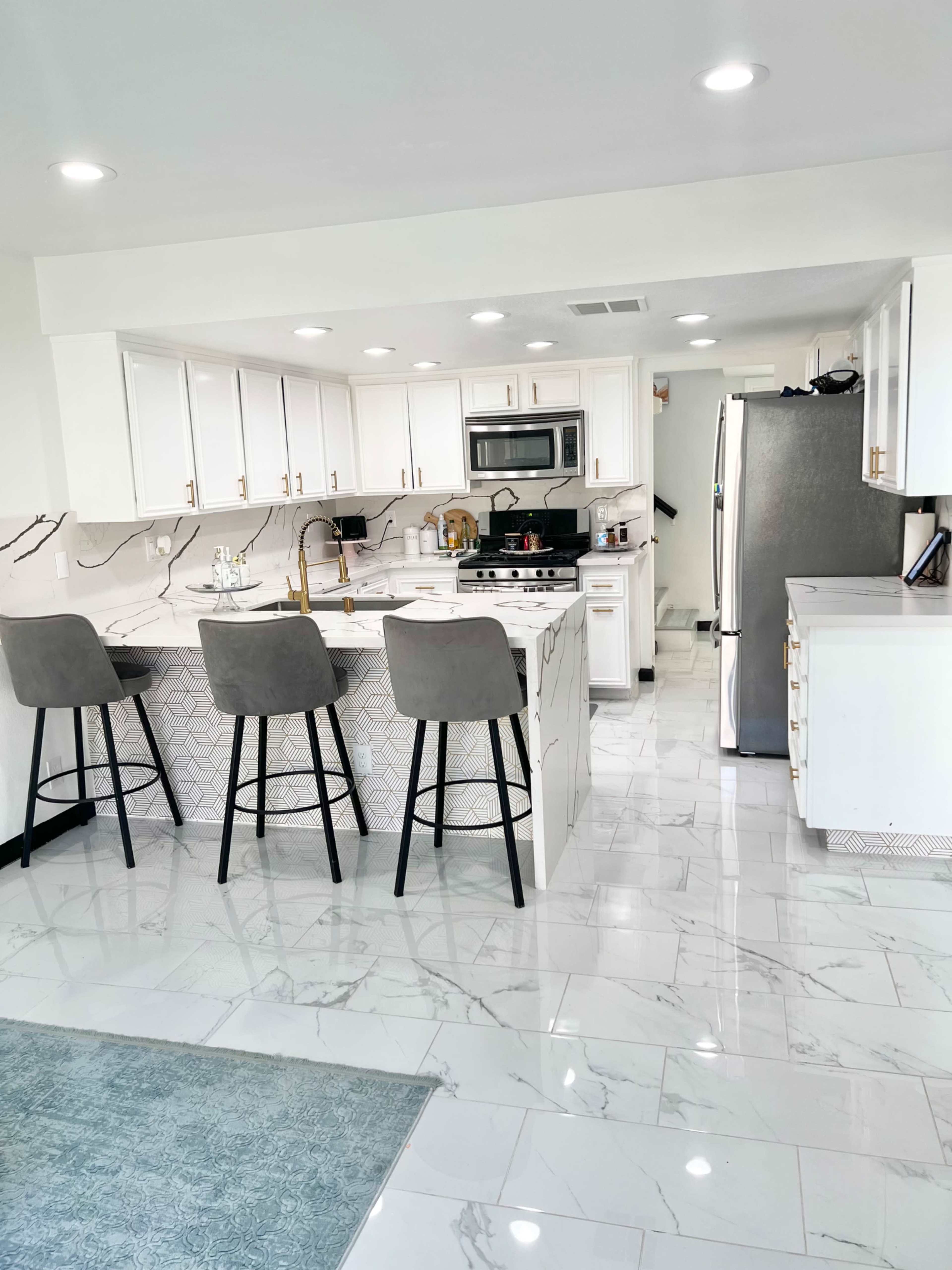 A modern kitchen features white cabinetry, marble countertops, stainless steel appliances, and a breakfast bar with three gray stools.