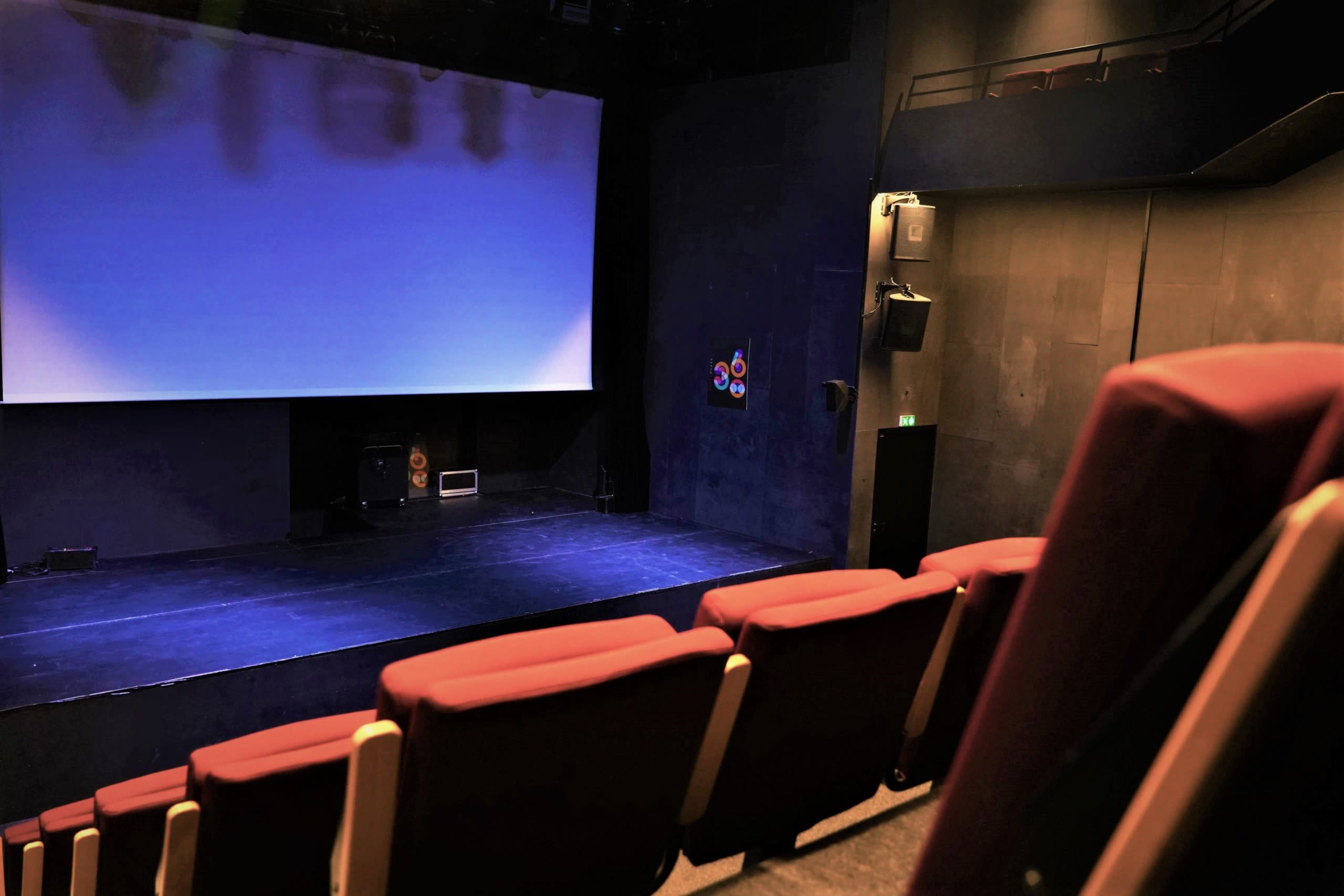 An empty theater with red seating and a dark stage illuminated by blue lighting.