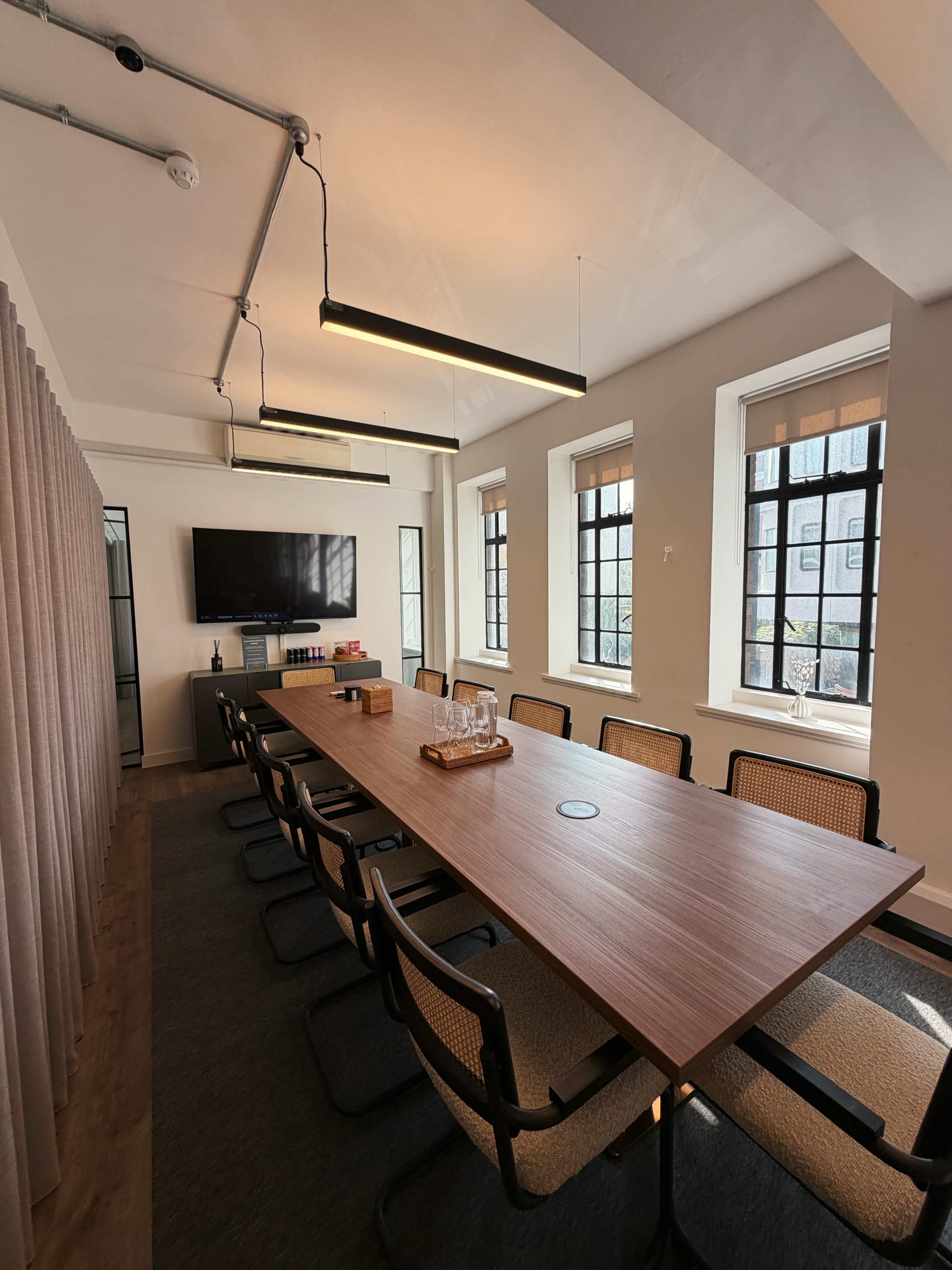 A modern conference room features a long wooden table surrounded by black chairs, with large windows providing natural light and a television mounted on the wall.