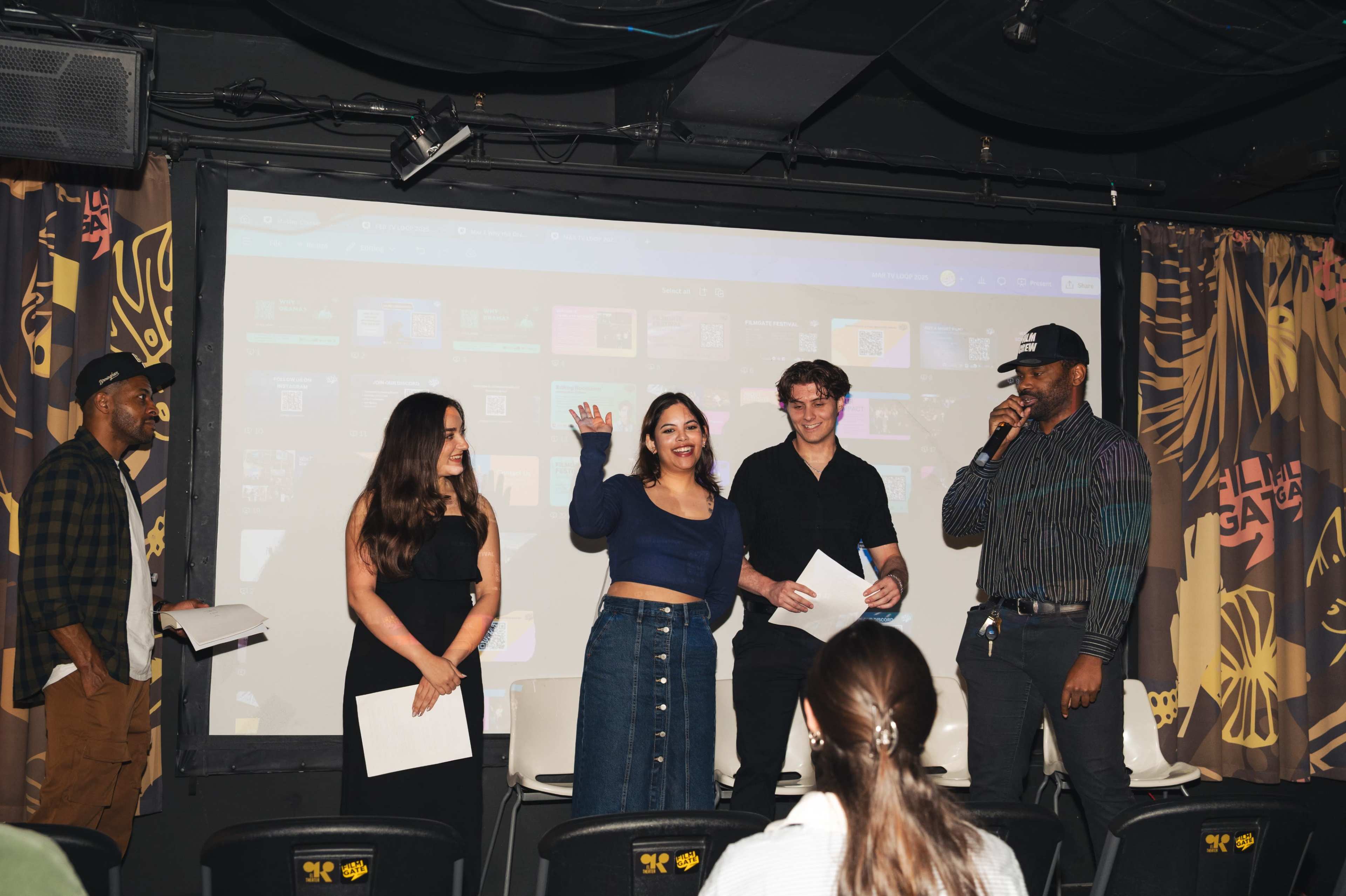 A group of four people stands on a stage in front of a large screen, engaging with an audience at an event.