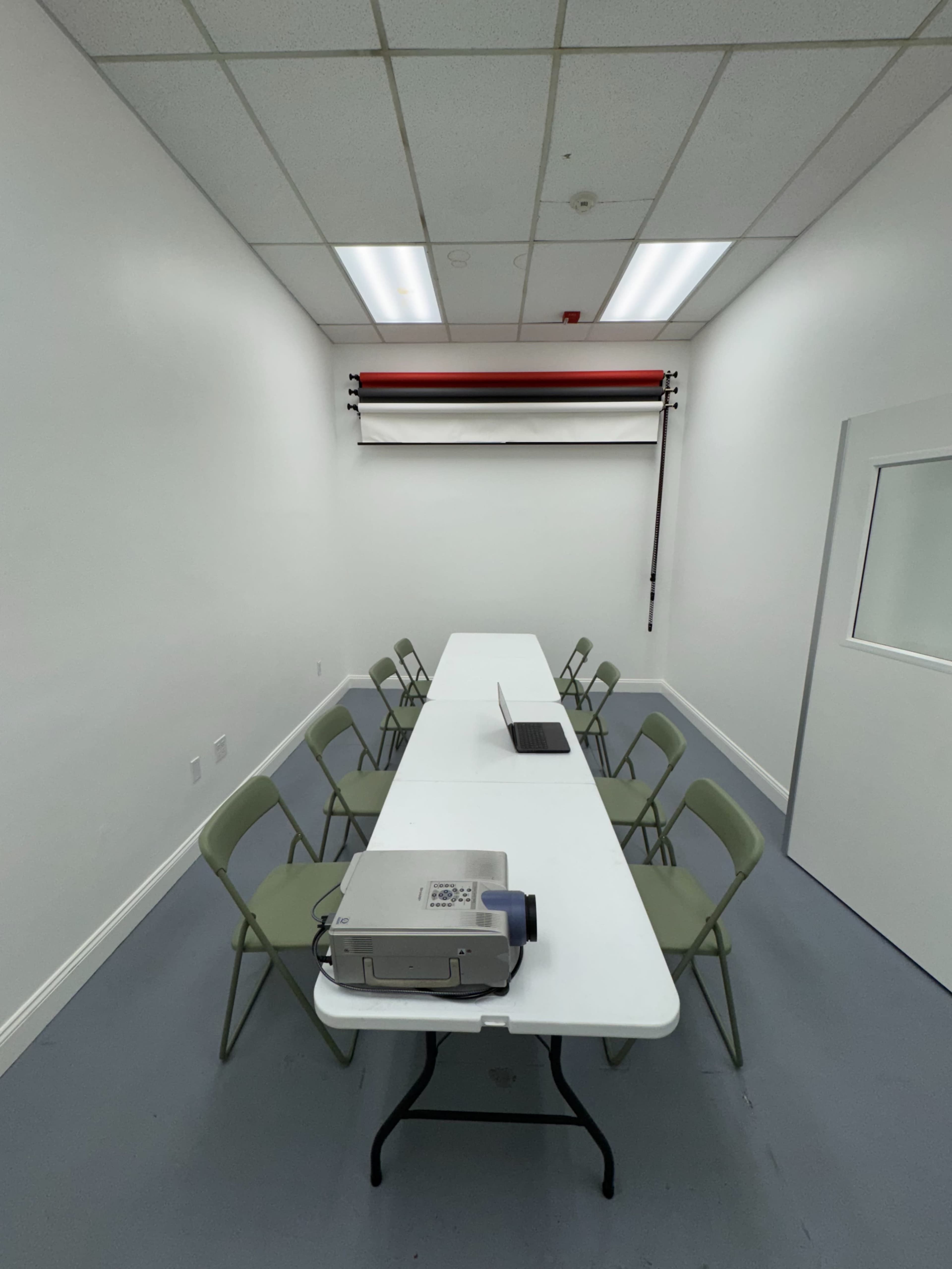 The image shows a small, empty meeting room with a long table, several green chairs, a laptop, and a projector on the table, with white walls and a roll-up screen at the far end.