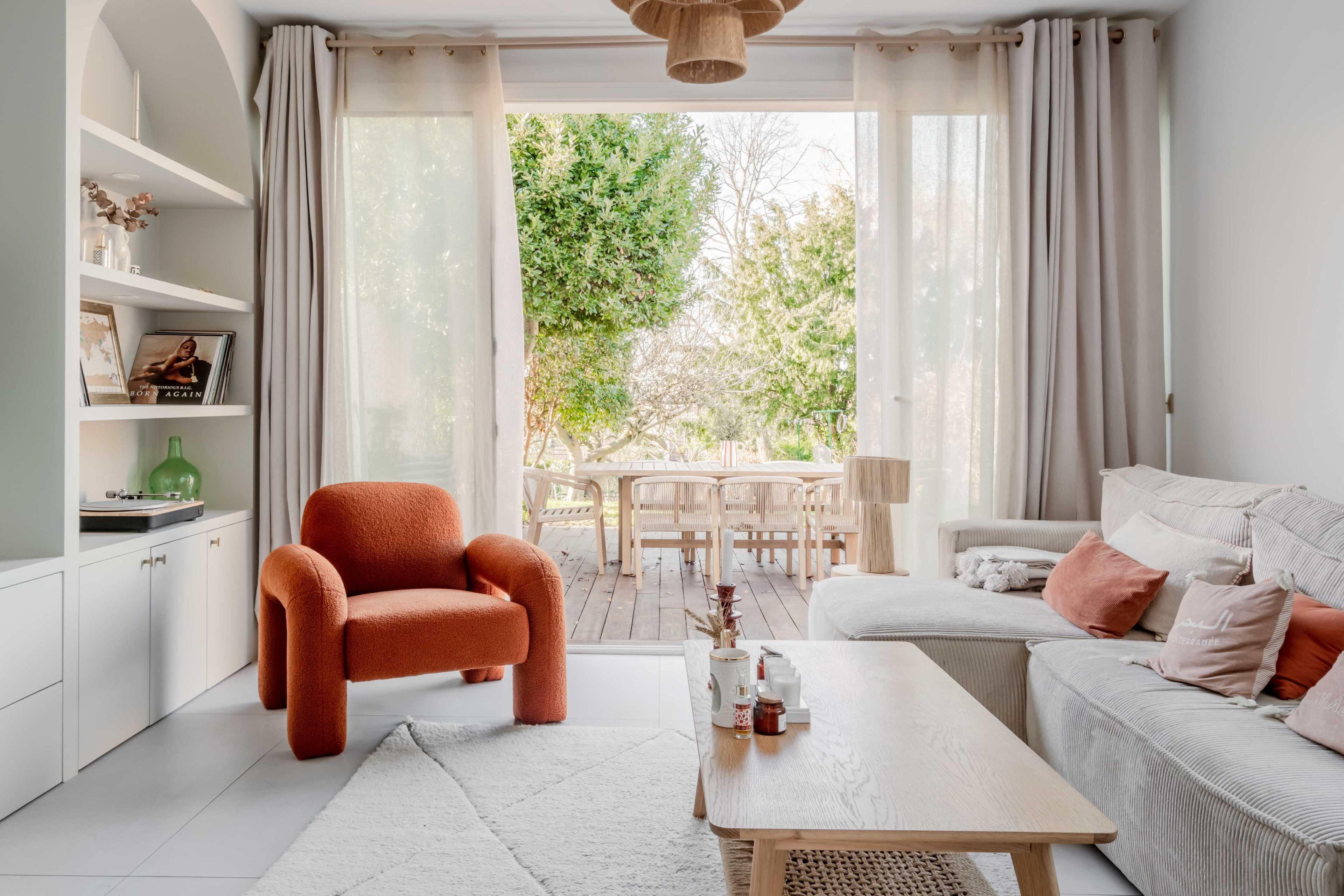 A cozy living room features a neutral sofa and an orange chair, with large windows that open to a wooden deck and a garden area.