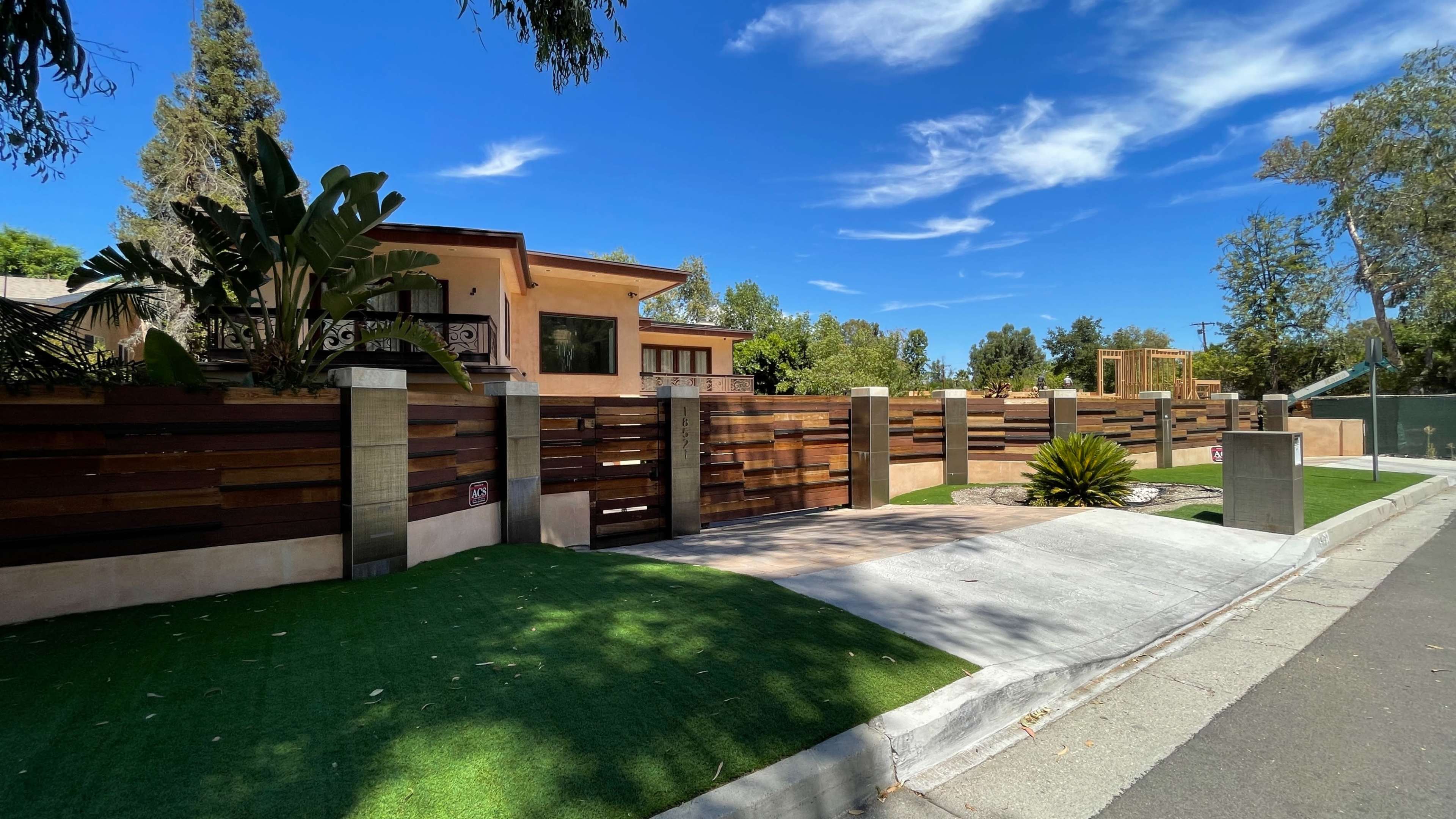 The image shows a modern home with a wooden fence and landscaped greenery along a quiet street.