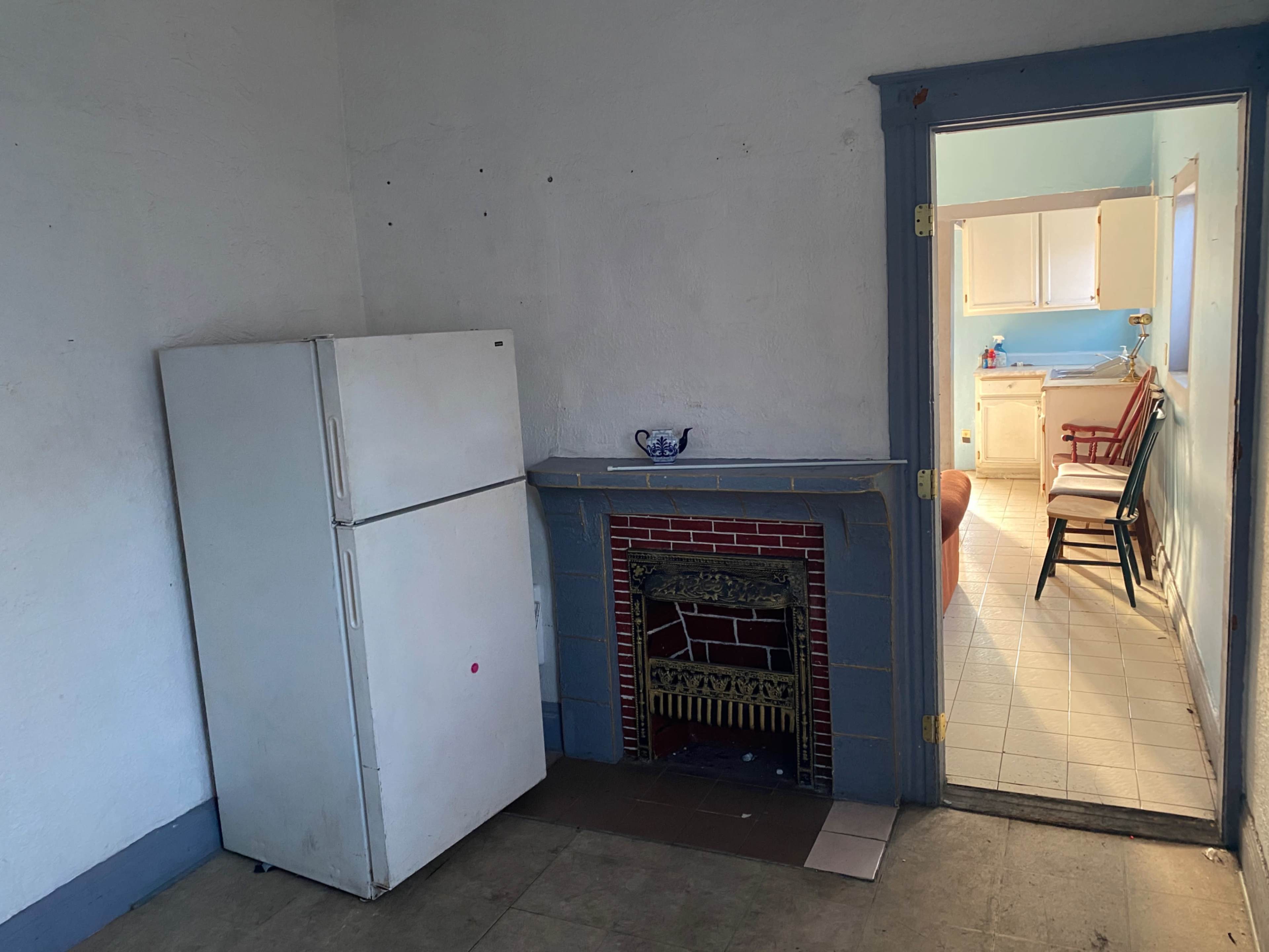 A white refrigerator stands next to a decorative fireplace in an empty room with an open doorway leading to a kitchen.