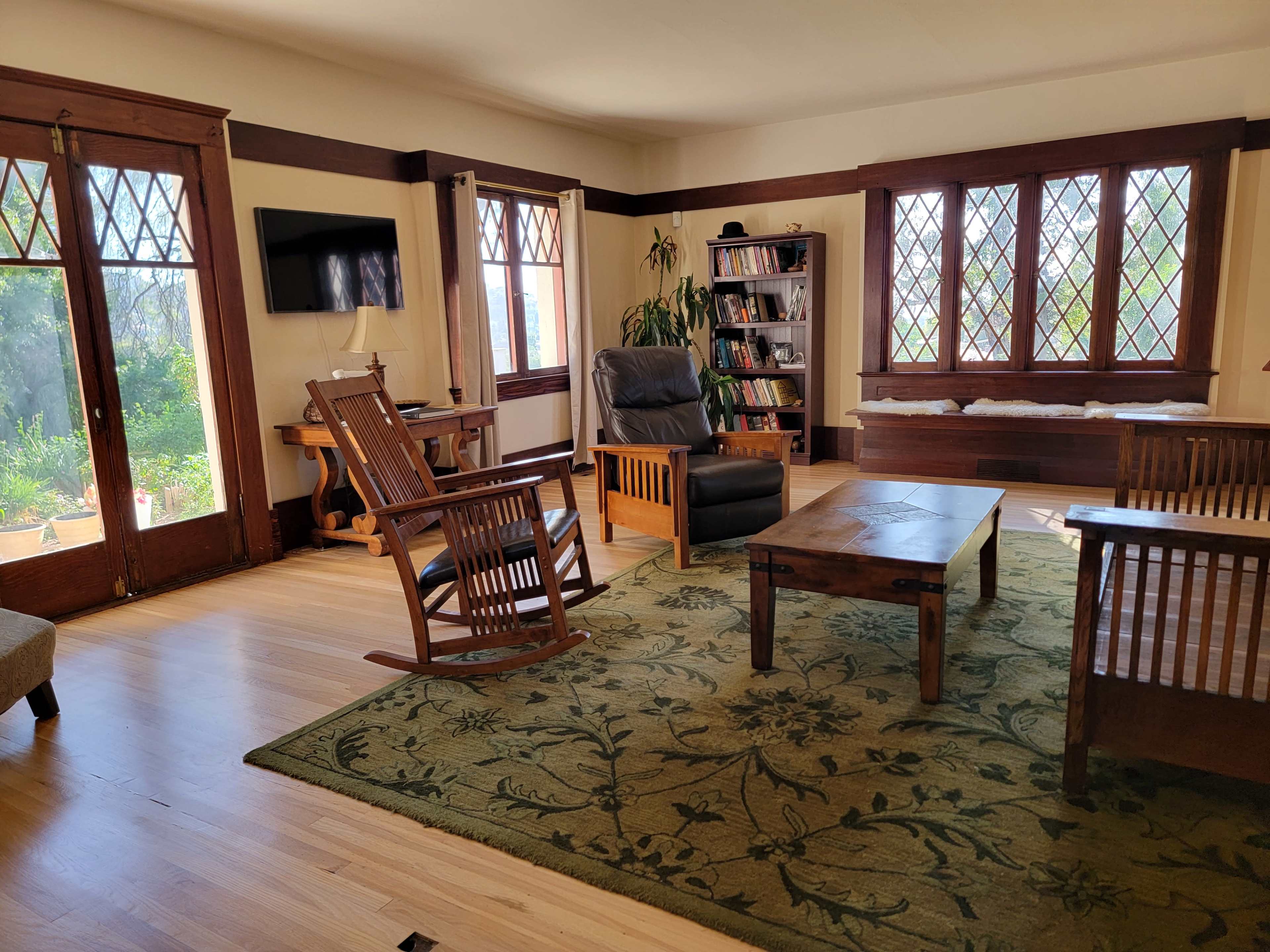 The image shows a bright living room with a wooden rocking chair, a leather recliner, a coffee table, and a bookcase, featuring large windows with diamond-shaped panes.