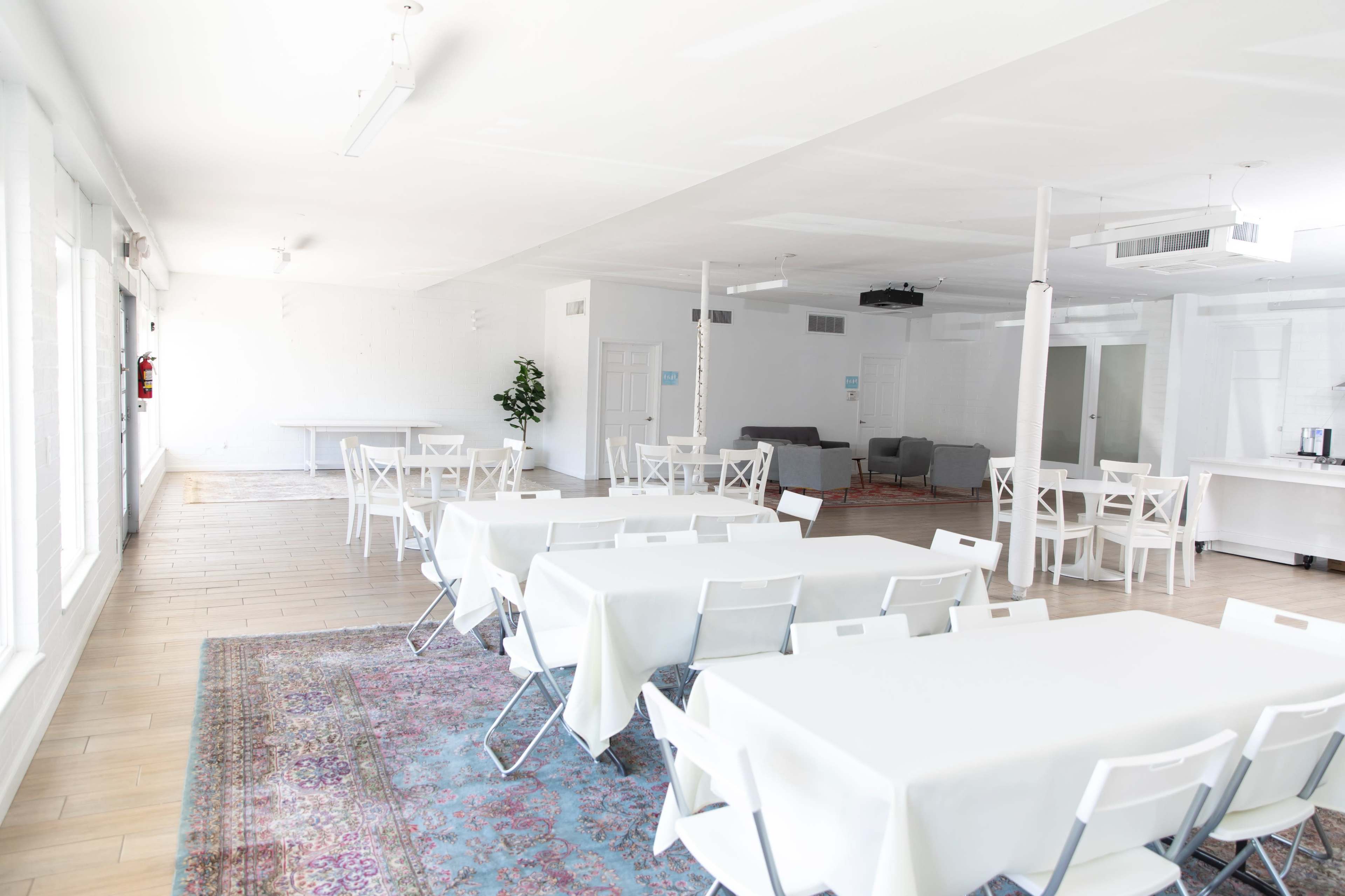 A bright, open event space with several white tables and chairs arranged on a patterned rug.