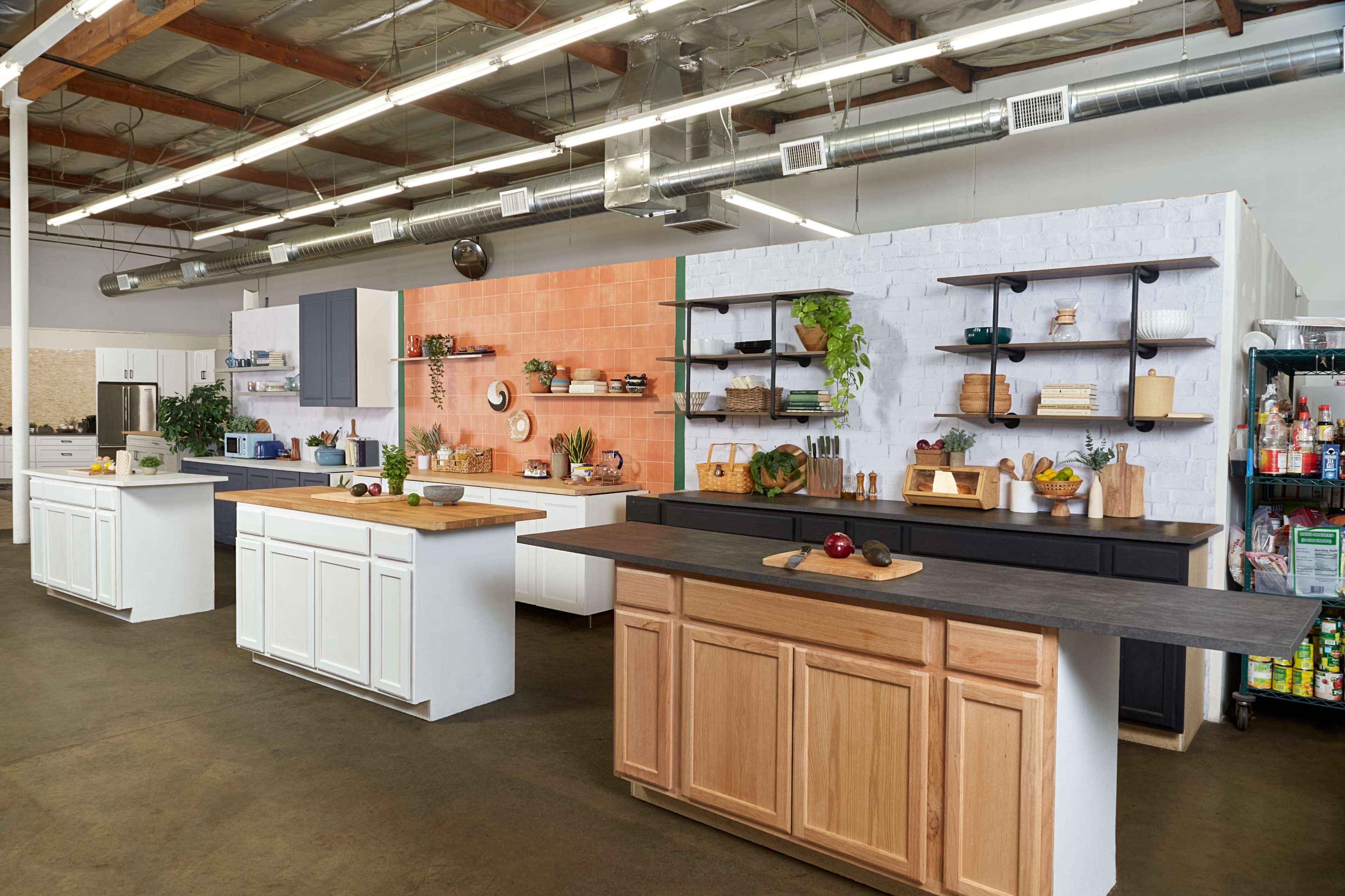 A spacious kitchen display area featuring multiple island counters and distinctive wall colors, with various kitchenware and plants arranged on shelves.