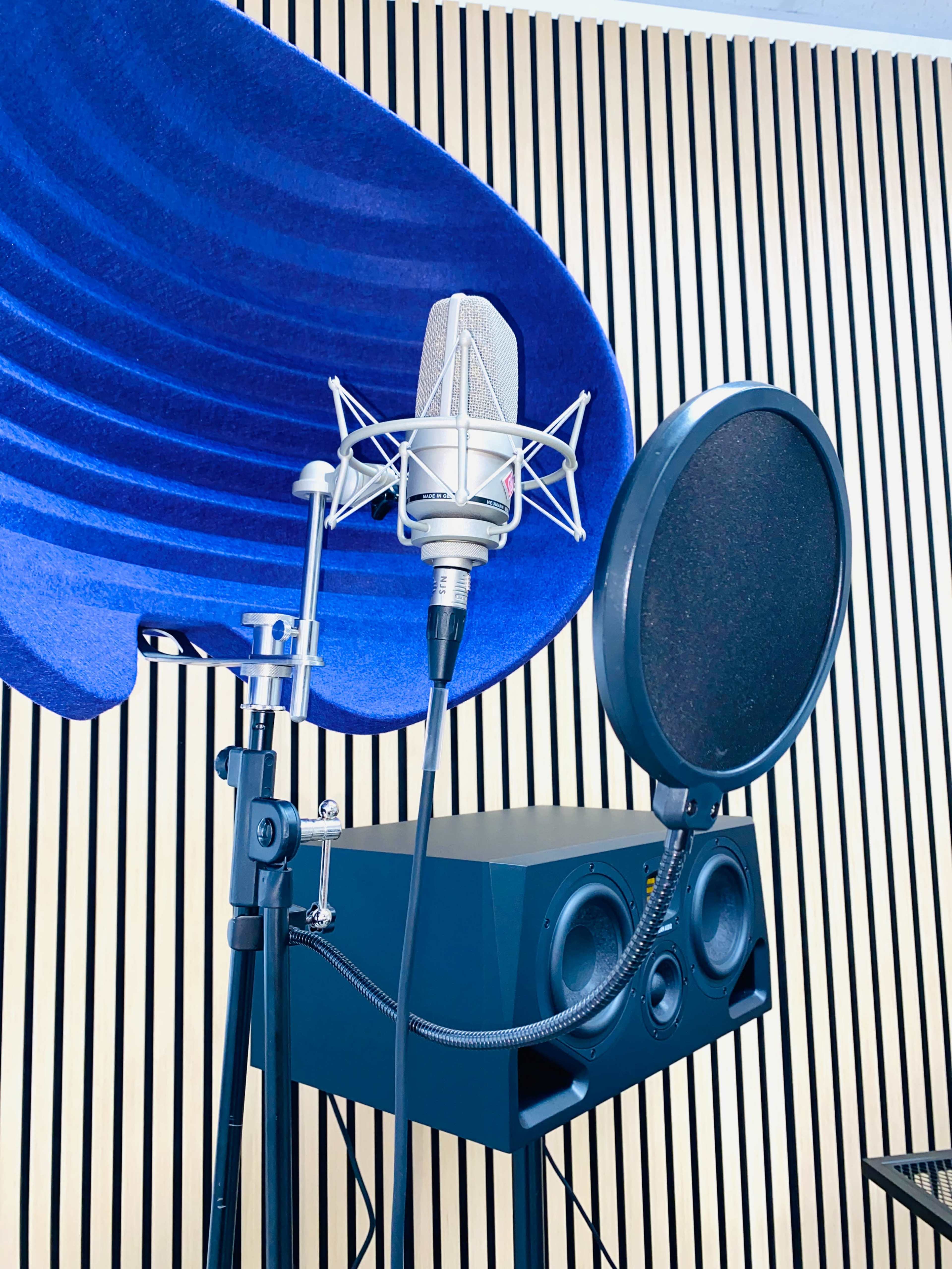 A microphone with a pop filter is mounted on a stand in front of a blue sound-absorbing panel, near a pair of speaker monitors.