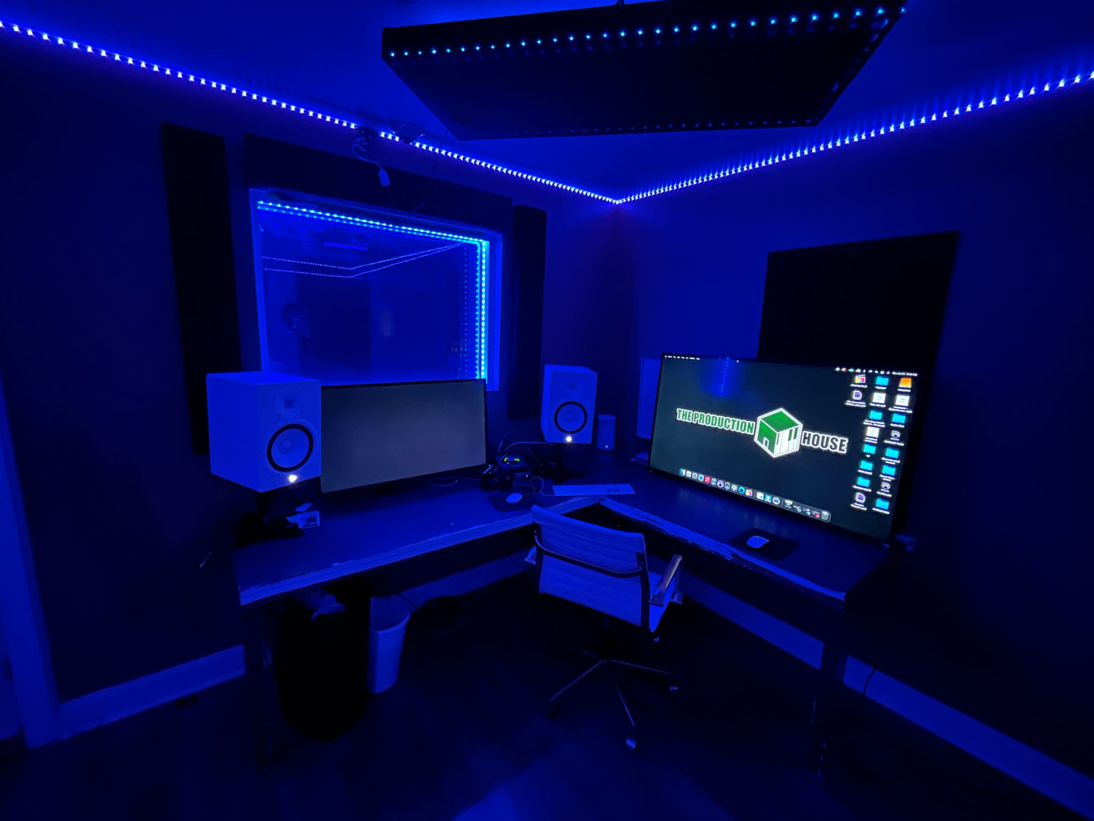 A dimly lit recording studio features blue LED strip lighting, two monitors, a pair of speakers, and a sleek desk with a rolling chair.