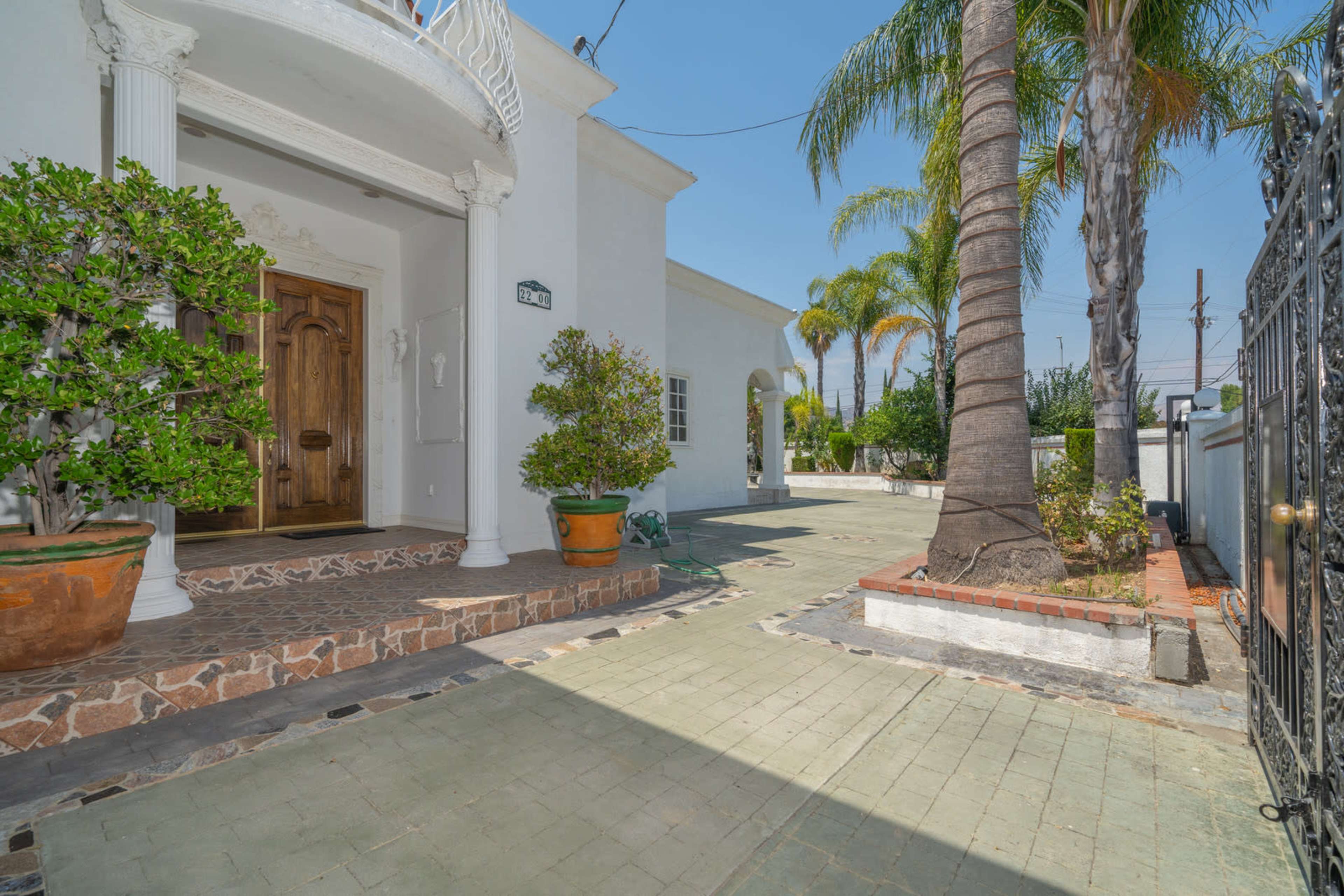 A white stucco house with a wooden front door, surrounded by potted plants and palm trees, on a paved driveway.