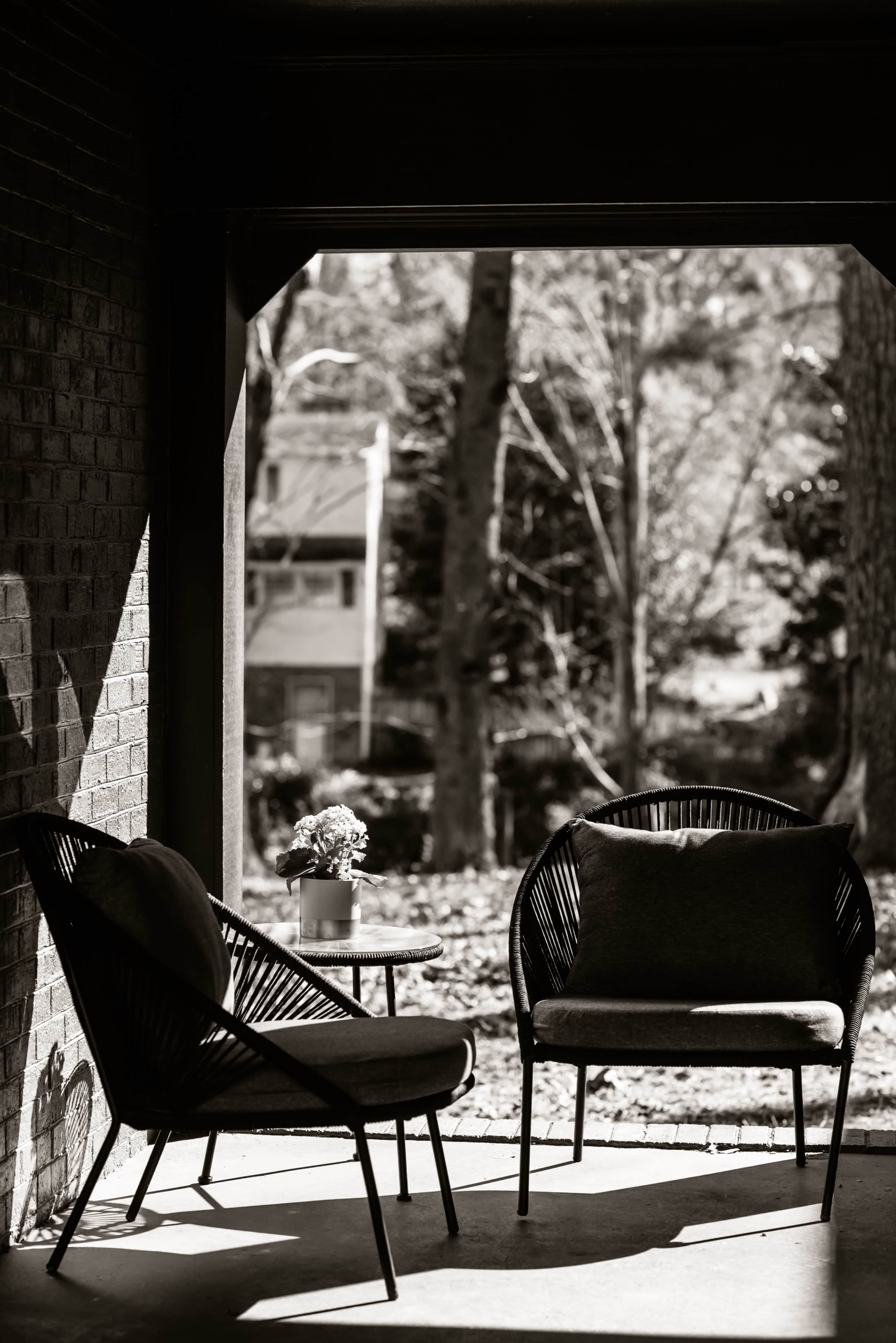 Two black rattan chairs and a small round table with a potted plant, framed by a shaded porch overlooking a wooded area.