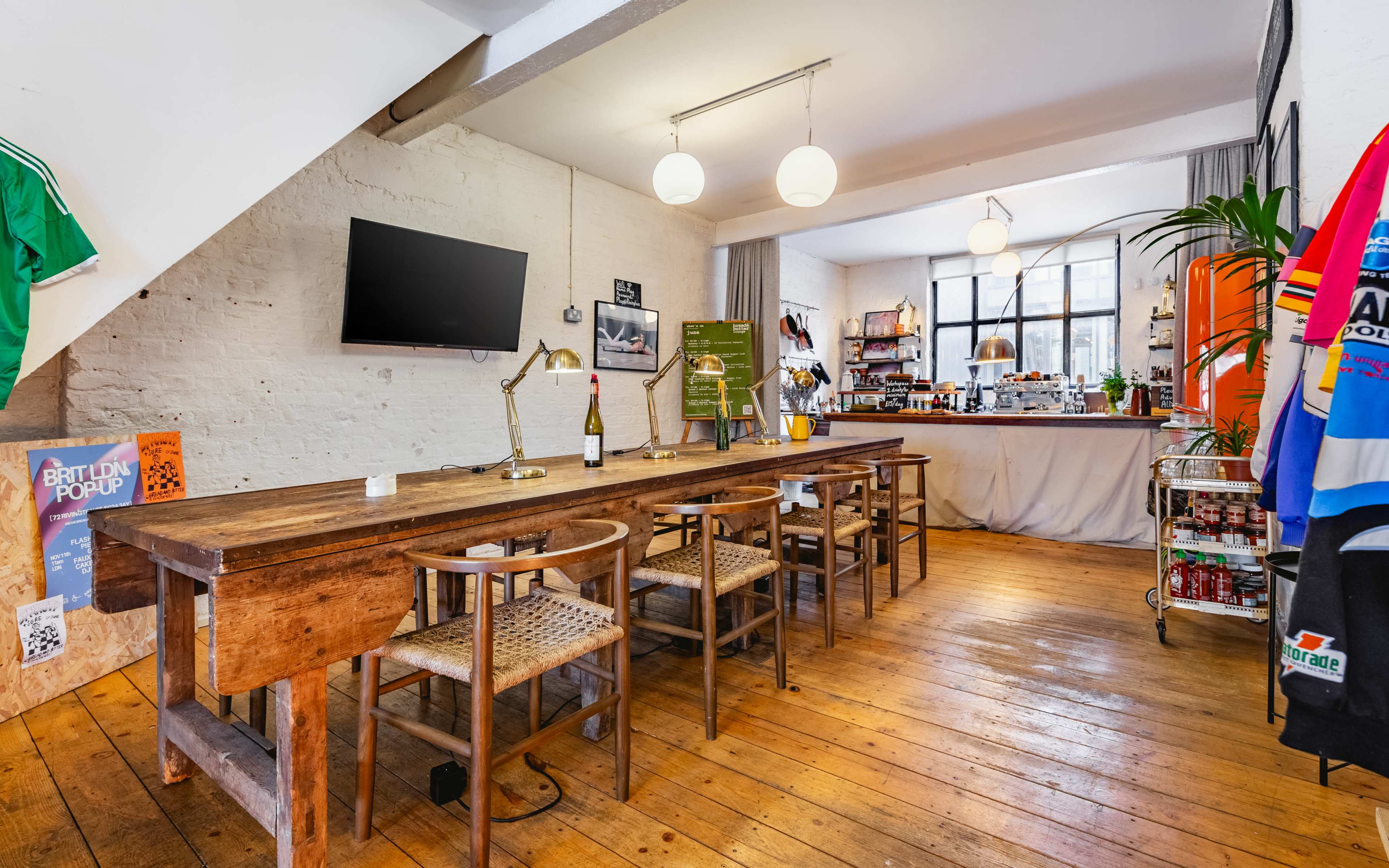 A long wooden table with several chairs is positioned in a brightly lit room featuring a kitchenette and a television on the wall.