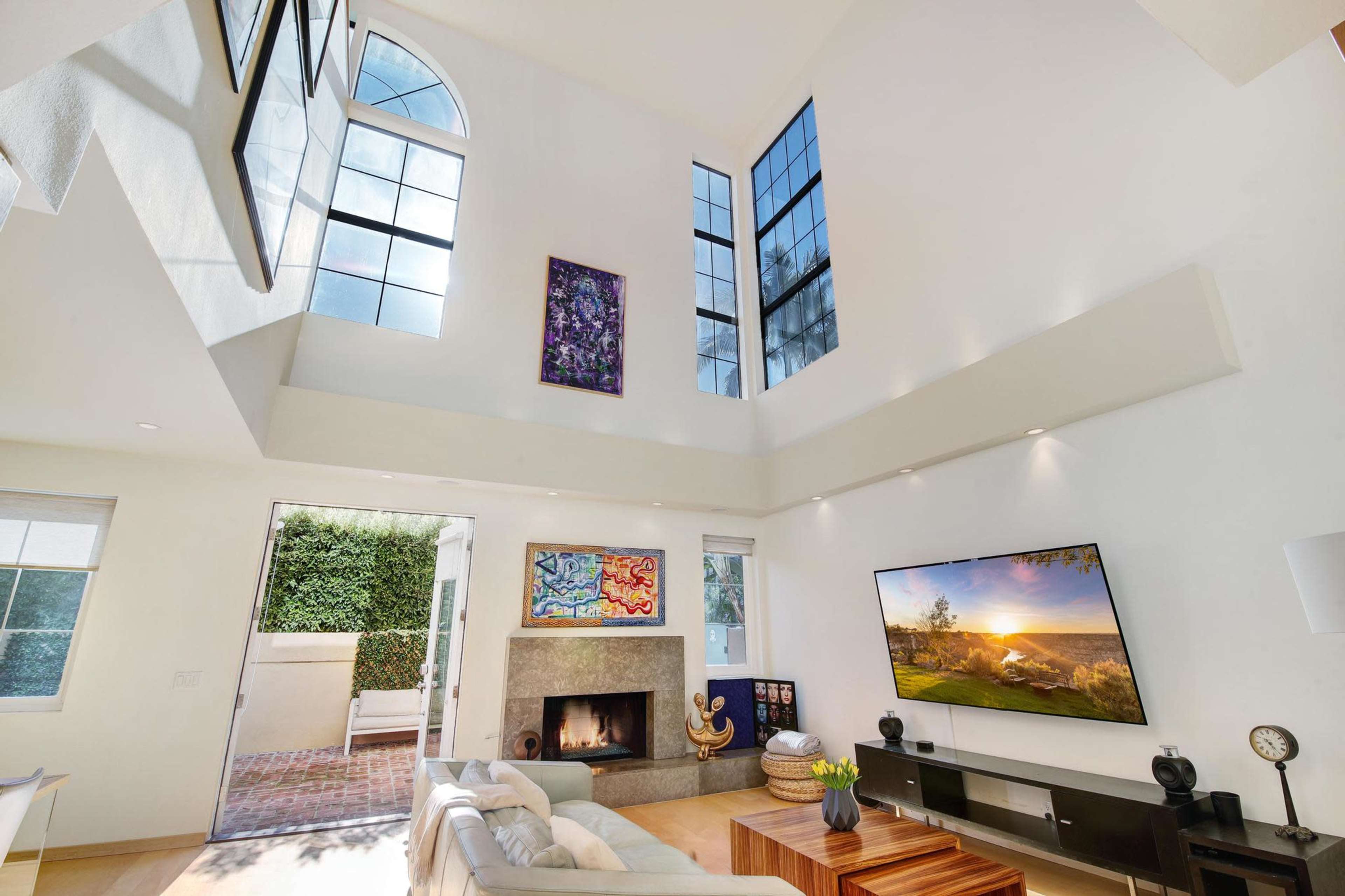 The bright living room features large windows, a mounted television, and a fireplace, with access to an outdoor patio.
