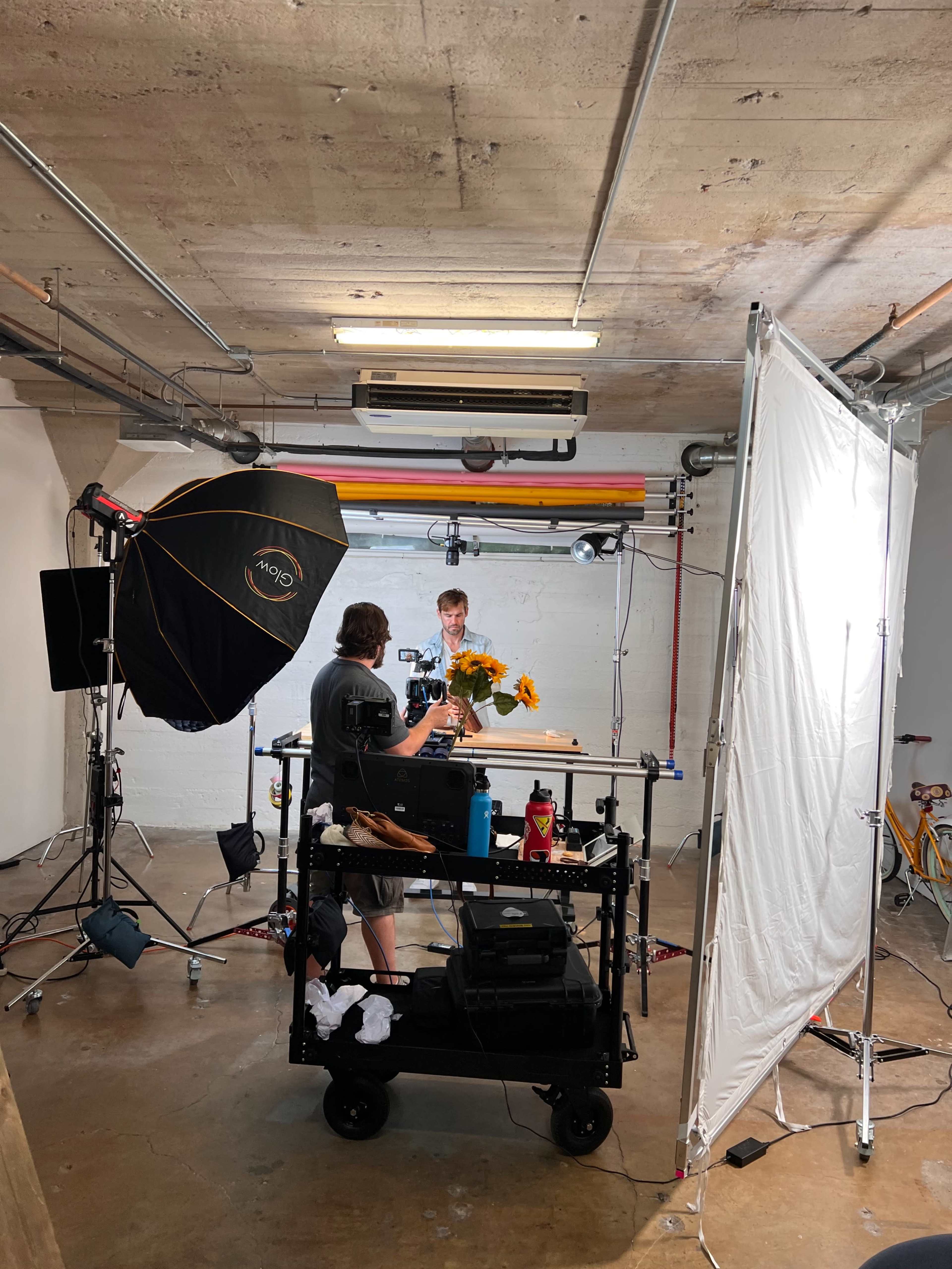 A photographer is setting up equipment and arranging flowers on a table in a studio with various lighting and a backdrop.